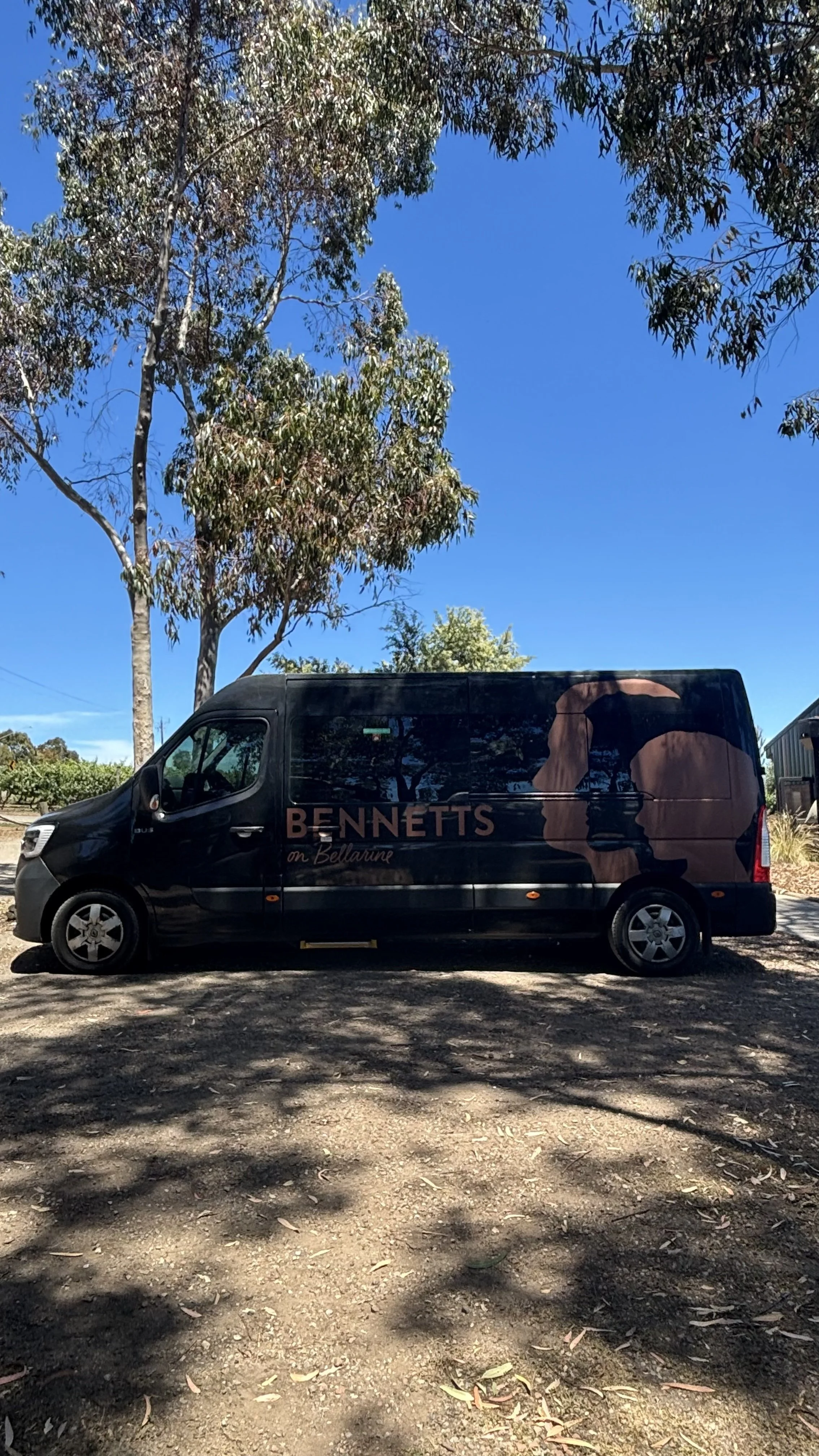 A black van with the words 'BENNETTS on Bellarine' and a silhouette of a woman's profile on the side, parked on a dirt area under a clear blue sky, with trees casting shadows nearby.