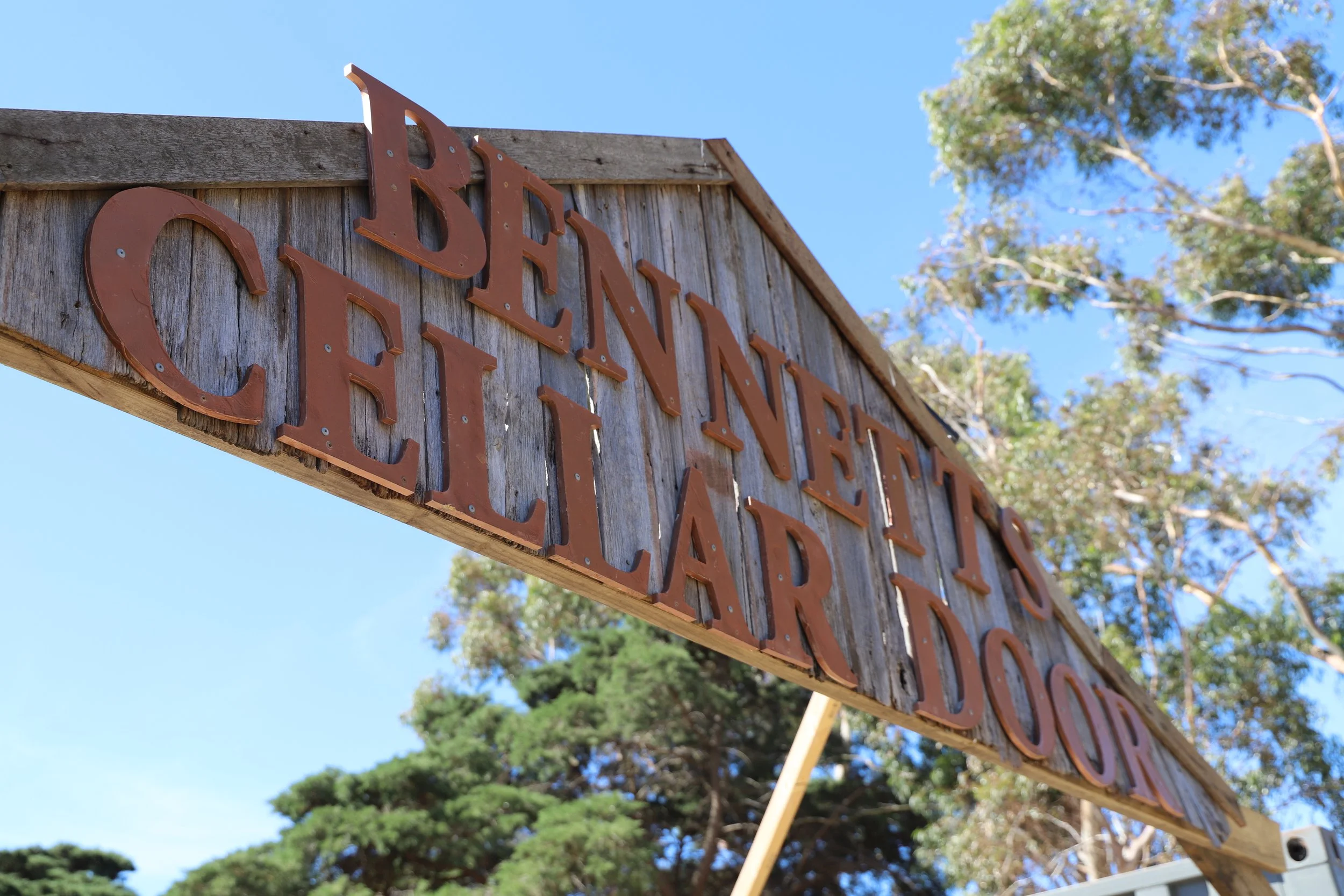 Wooden sign with red metal letters that reads 'BRENTAN PORT CALIFARDO' against a blue sky with trees in the background.