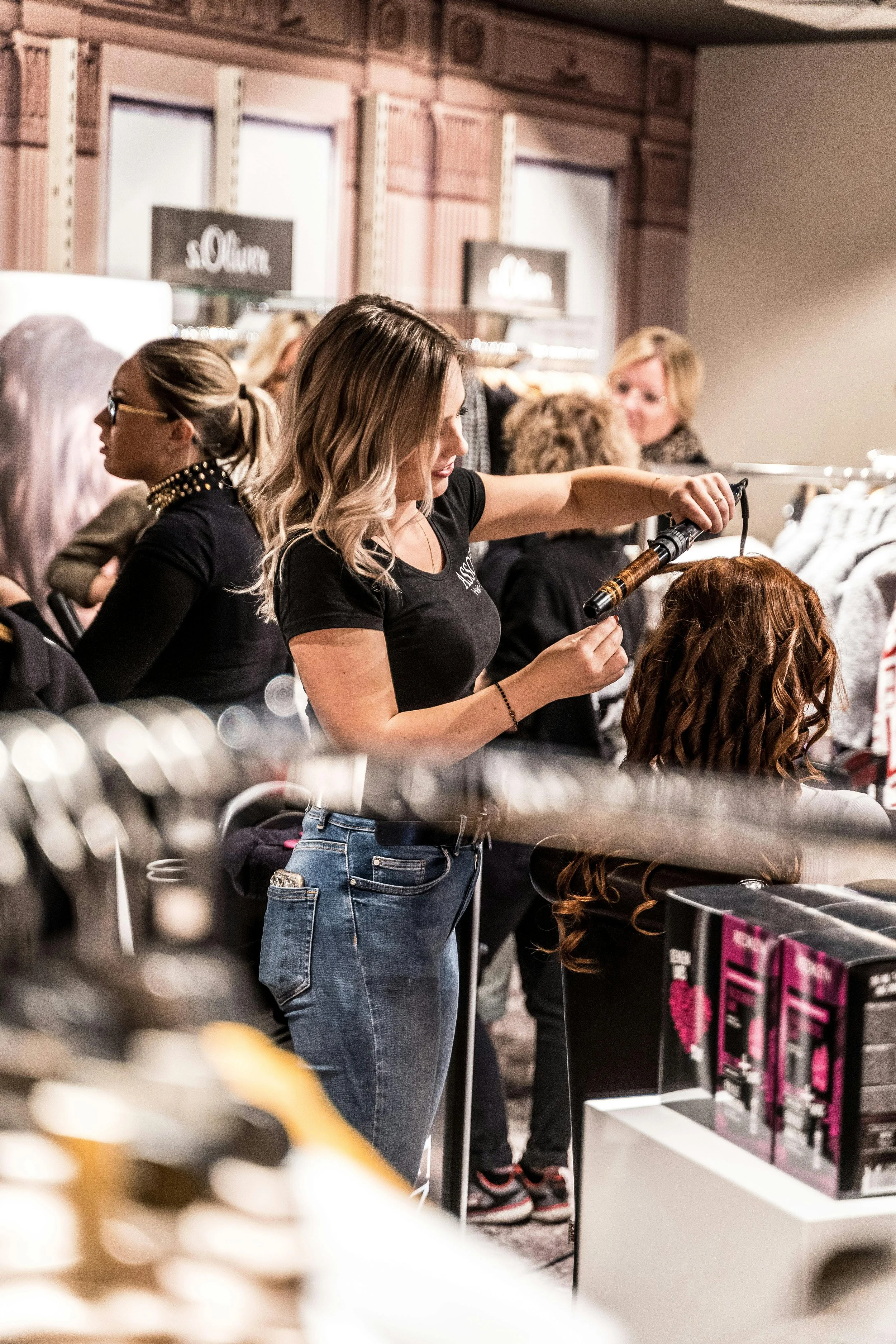 A woman with blonde hair curling a wig at a store, surrounded by clothing racks and shoppers.