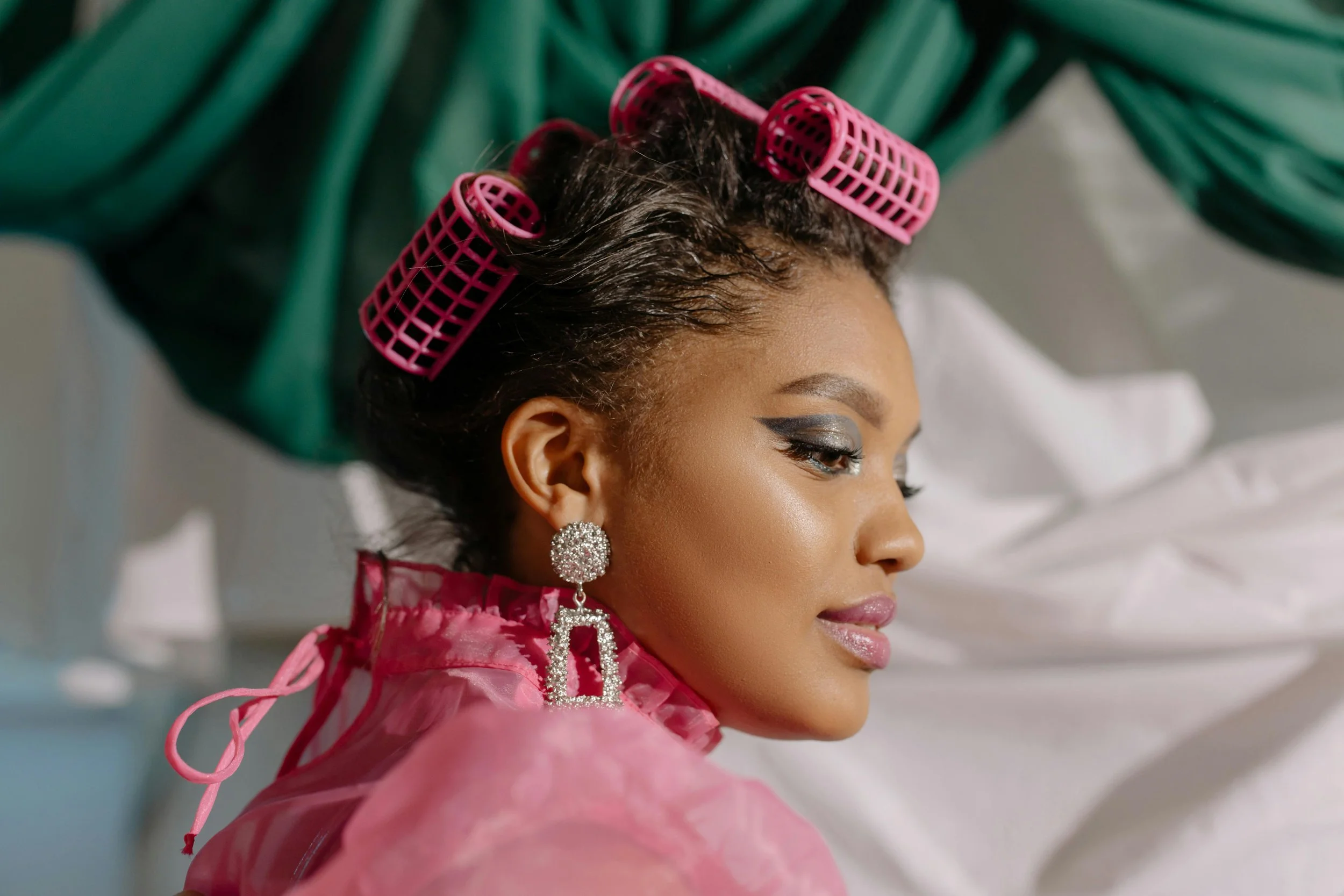 A woman with hair rollers in her hair, wearing pink clothing and large diamond earrings, is sitting with her eyes closed, possibly getting ready for an event.