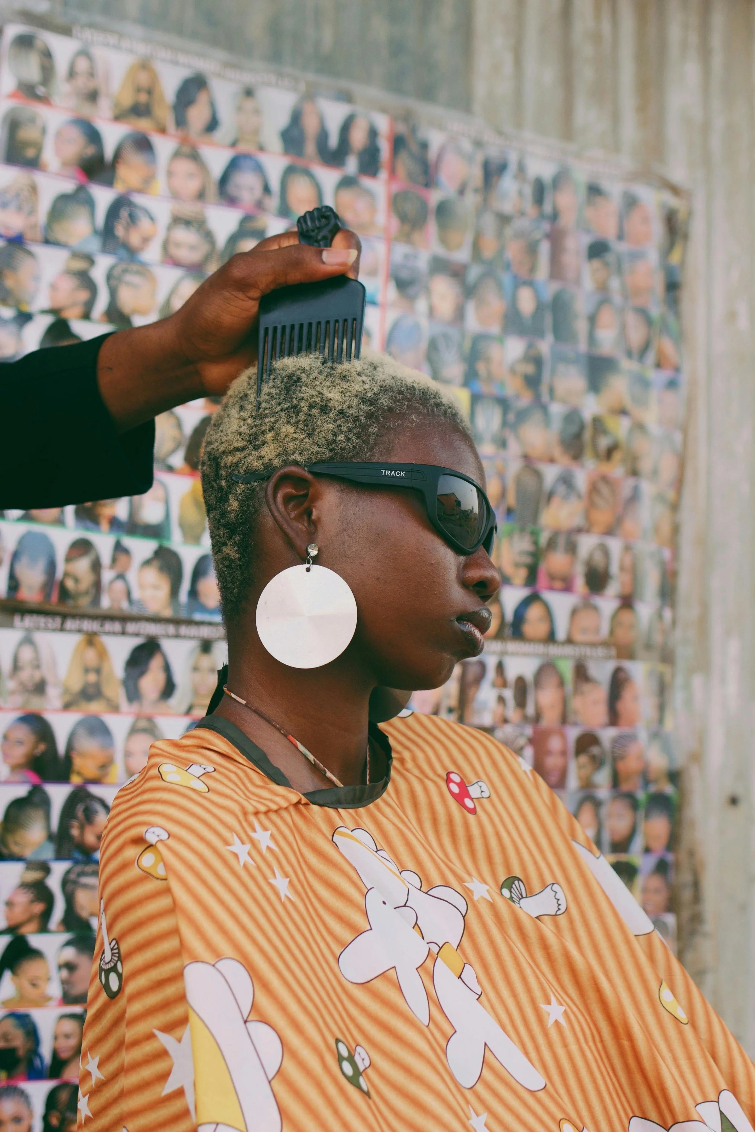 A woman with short, bleached blonde hair, large white earrings, and futuristic sunglasses, sits while a hairstylist combs her hair, with a collage of various hairstyles on the wall behind her.