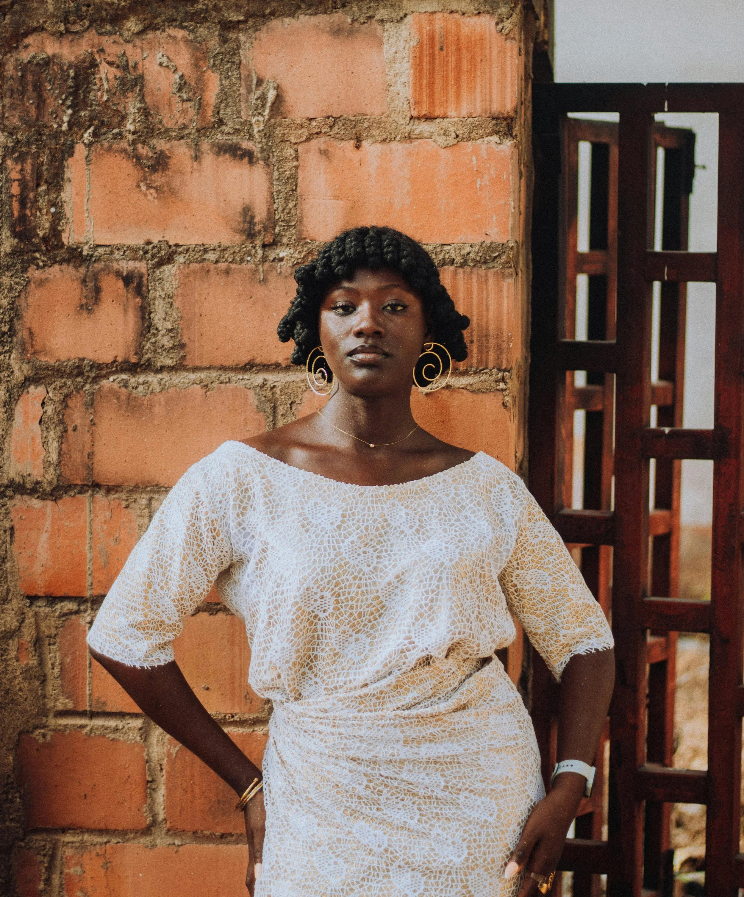 A woman standing against a brick wall and wooden gate, wearing a white lace dress, gold earrings, and a wristwatch, looking at the camera.