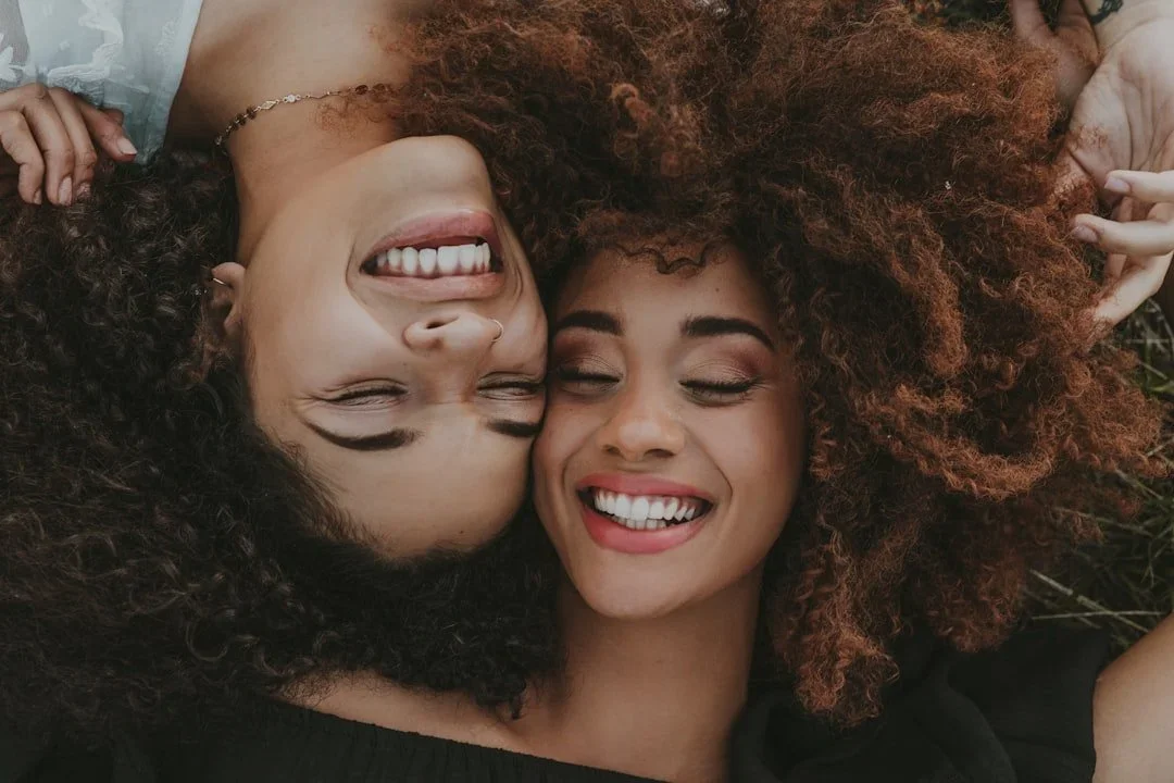 Two women with curly hair lying close together on grass, smiling with eyes closed.