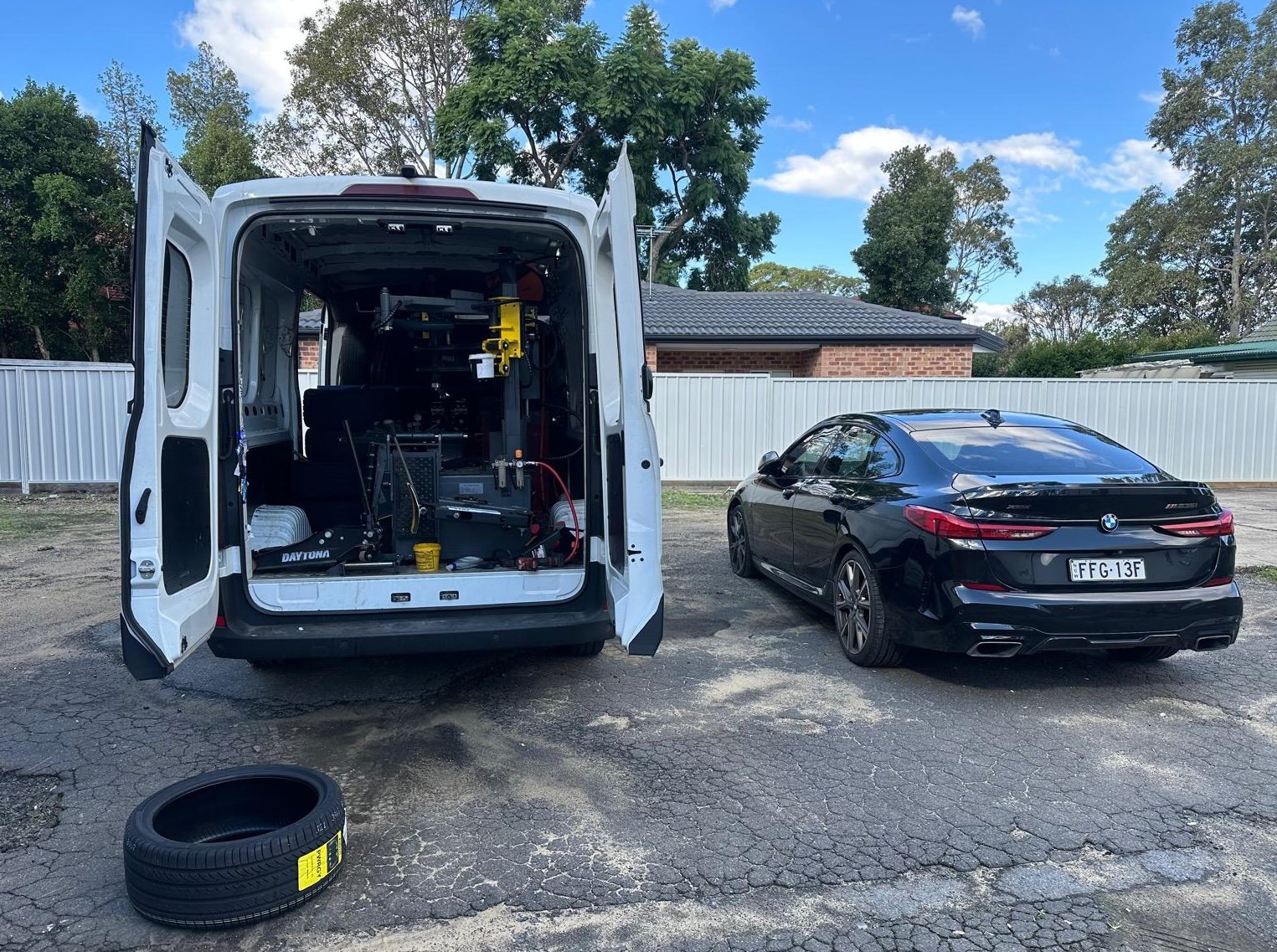 A white work van with its back doors open, showing equipment inside, parked on a cracked asphalt surface next to a black BMW coupe, with a loose tire on the ground nearby, in a residential area with trees, a brick house, and a white fence in the background.