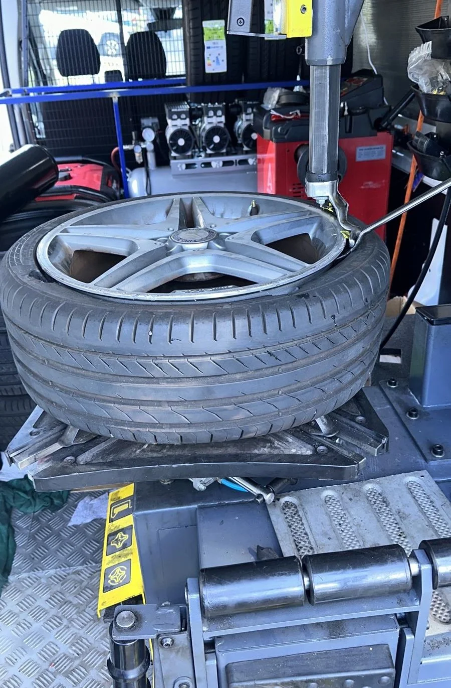 A wheel with a tire mounted on a machine in a workshop, likely for tire balancing or repair, with tools and equipment in the background.