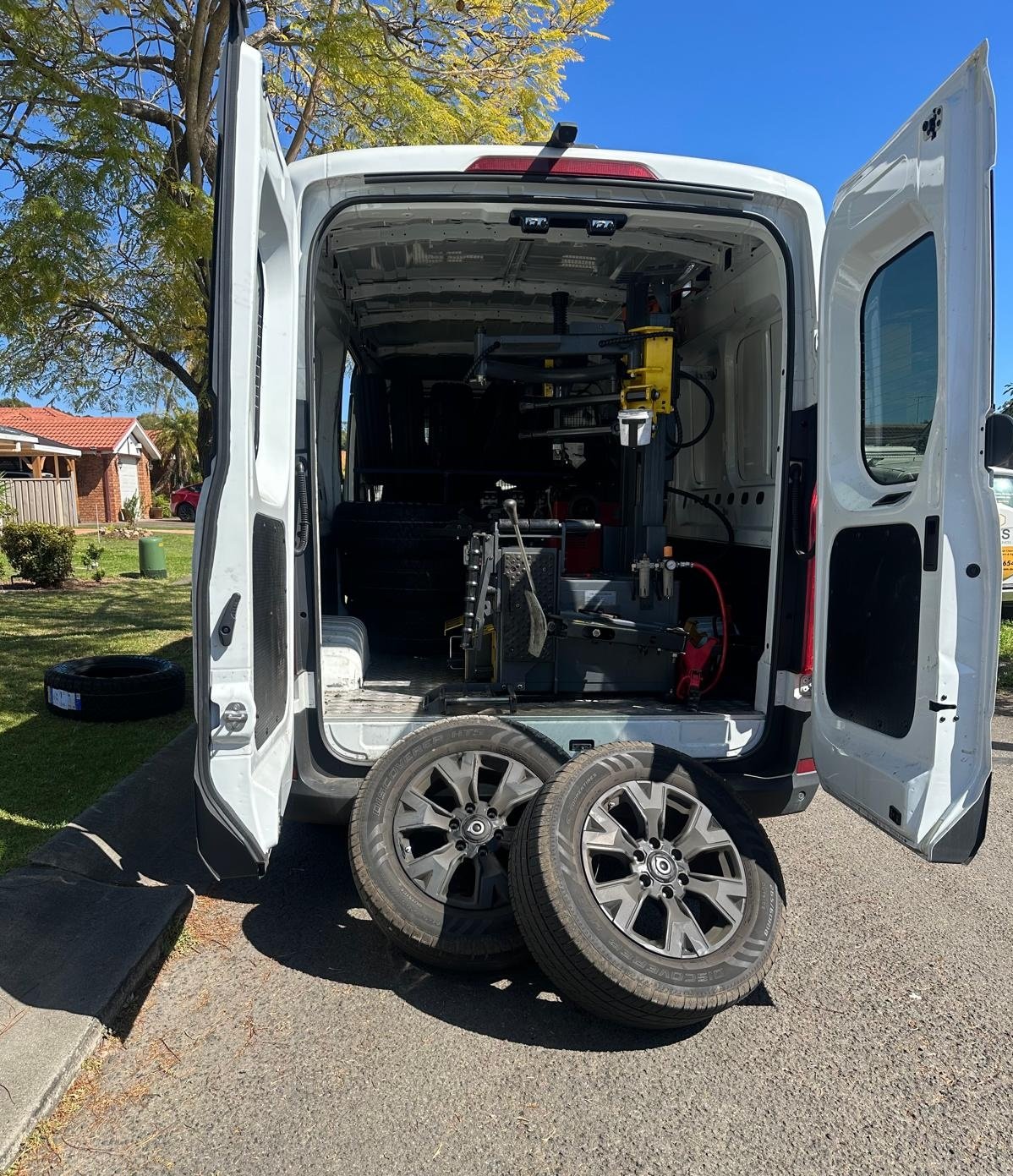 Back of a white cargo van with its rear doors open, showing equipment inside, with two tires leaning against the rear of the van, on a neighborhood street.