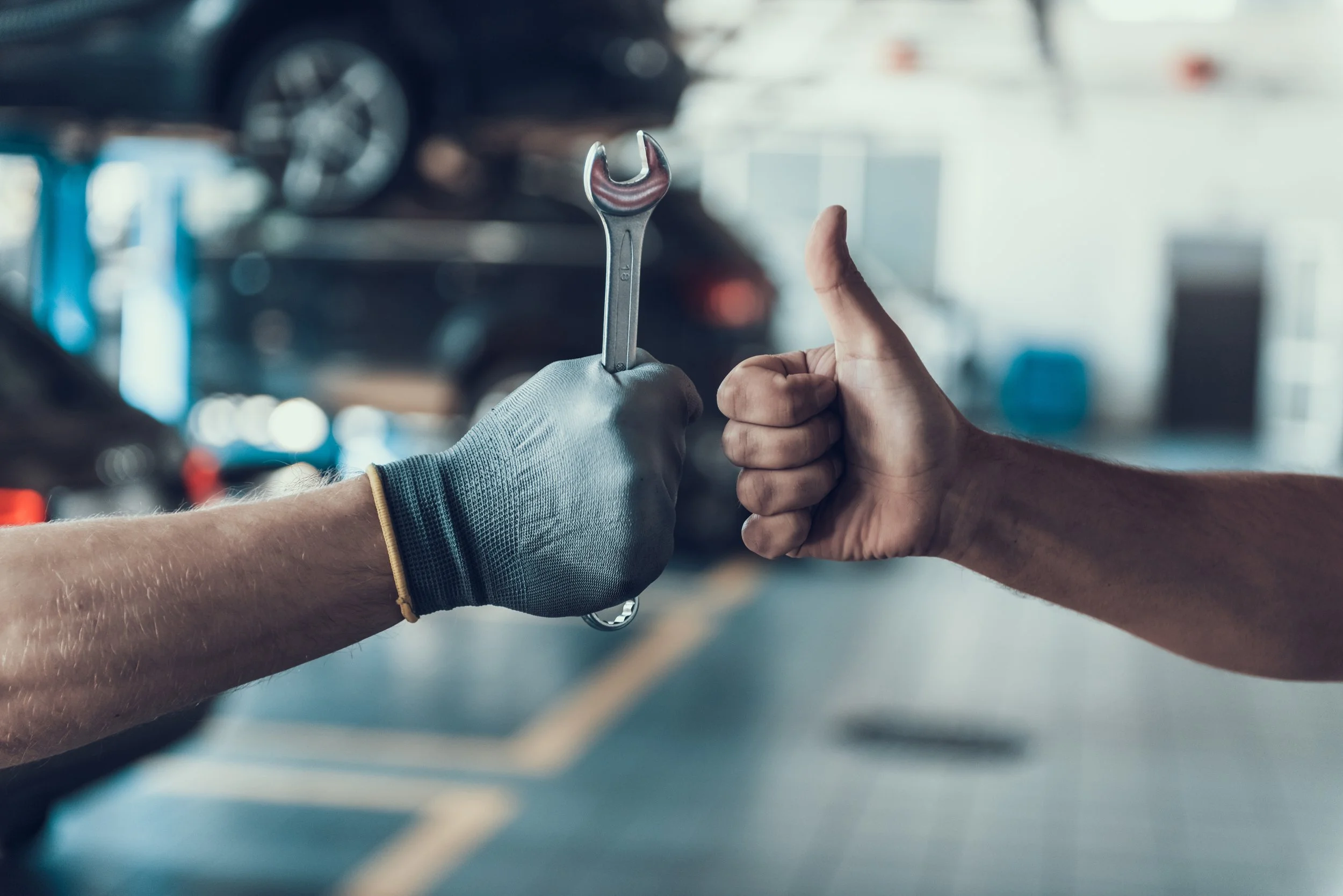 Person wearing a glove holding a wrench, while another person is giving a thumbs-up inside a car repair shop or garage.