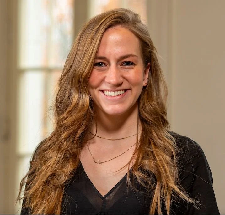 A young woman with long wavy blonde hair smiling, wearing a black top and layered necklaces, in front of a window with blurred background.
