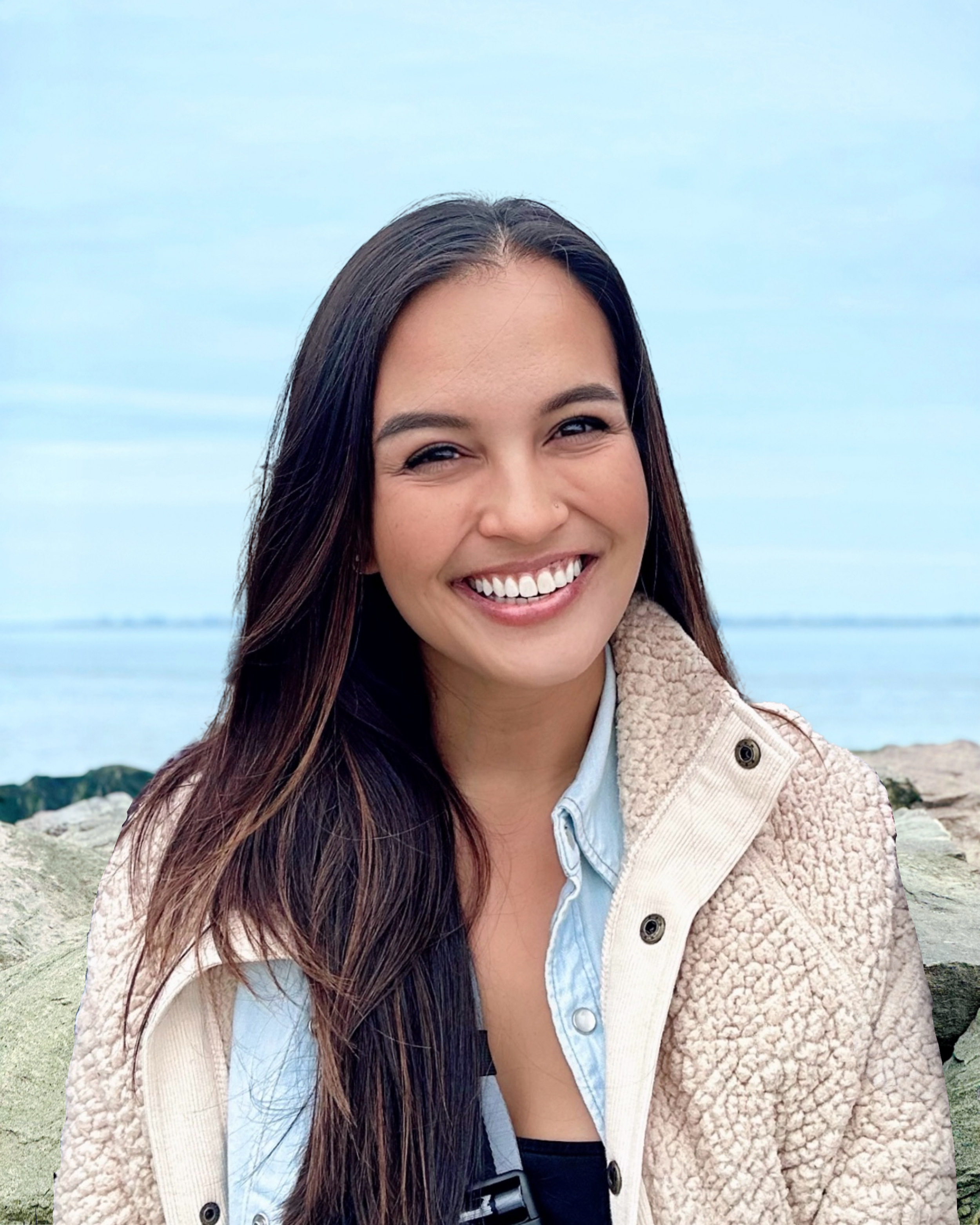 Rochelle Adriano, a smiling woman with long dark hair standing outdoors near water and rocks during daytime.