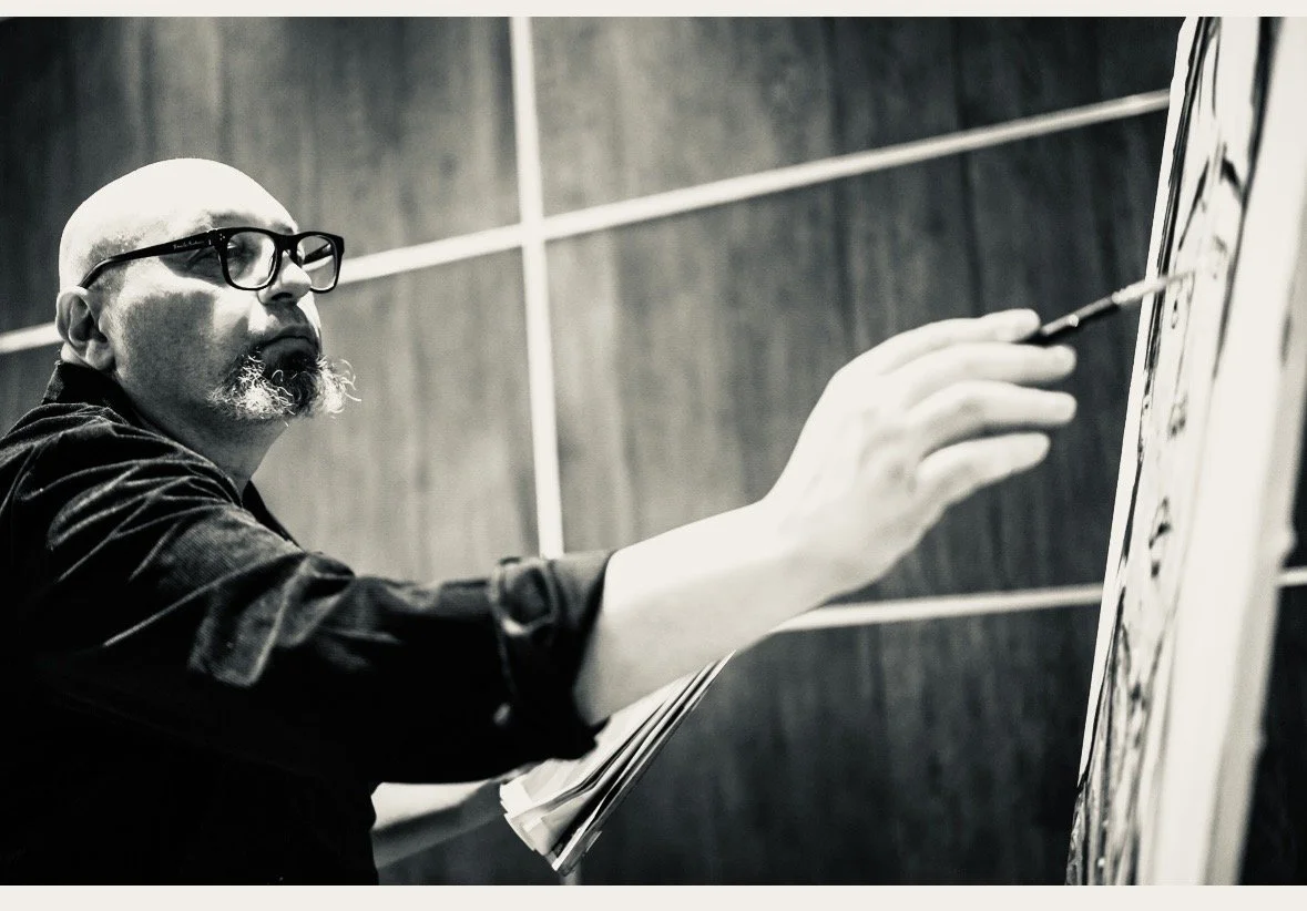 Artist with glasses and a beard painting on a canvas in a room with wood-paneled walls.