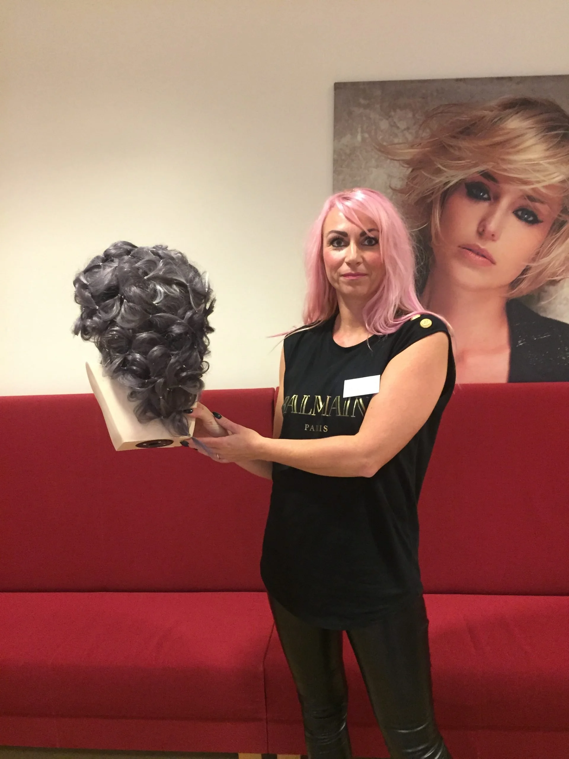 Kvindelig frisør holder en sort krøllet hårparykstander, står foran en rød sofa og et portræt af en kvinde med blåt øje makeup.