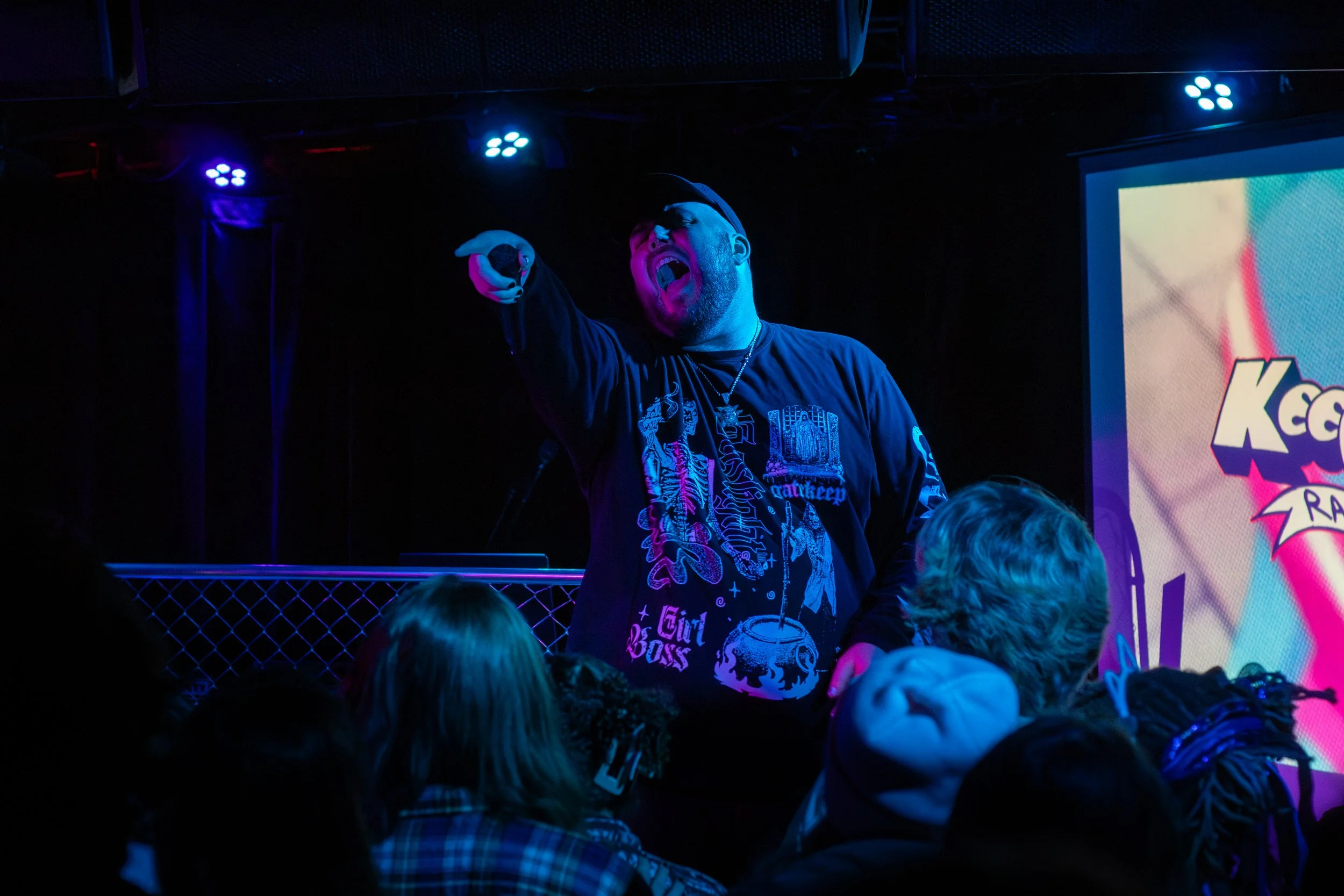 Performer singing or speaking on stage at a music event, pointing with his right hand, wearing a black cap and a black long-sleeve shirt with colorful graphics, surrounded by audience members in a dark venue with stage lights and a screen displaying 