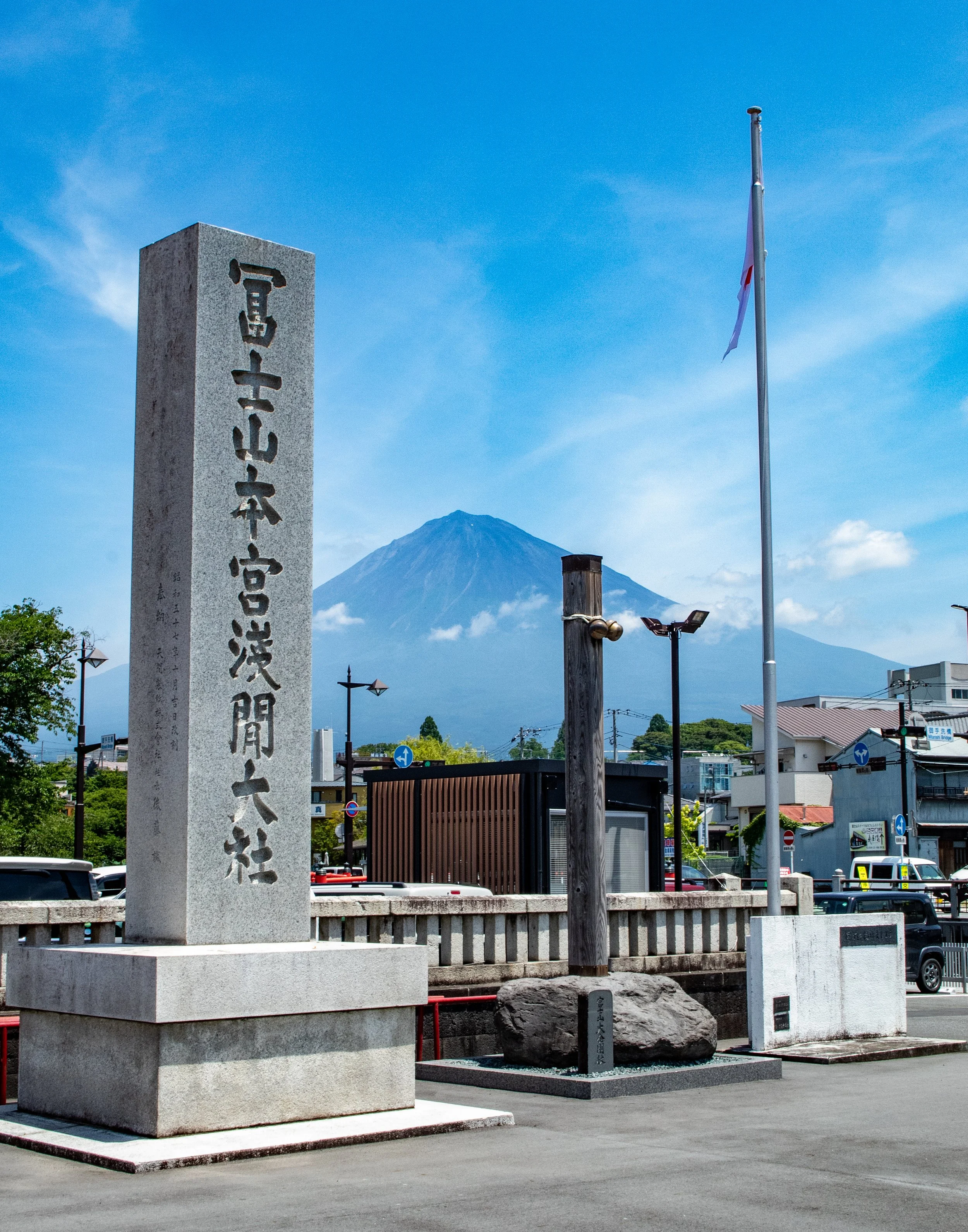 Statue with Japanese inscriptions, Mount Fuji in the background, flagpole with Japanese flag, urban area with buildings and cars.
