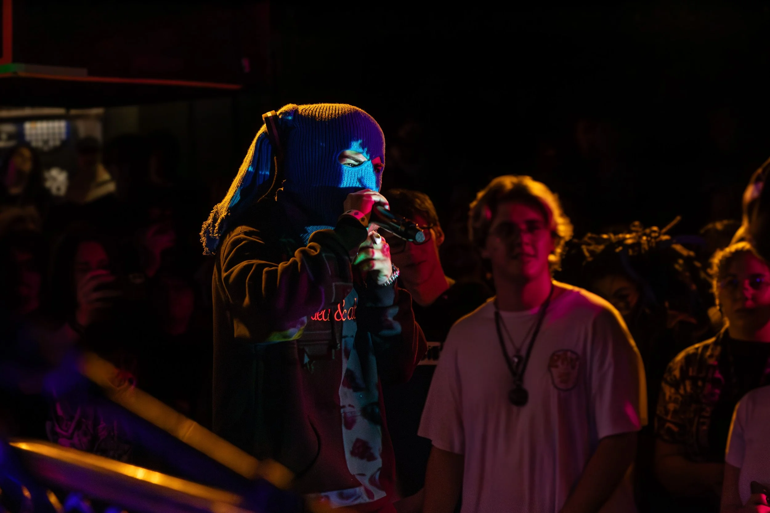 Person wearing a blue ski mask and black jacket holding a microphone, performing on stage with colorful lighting; audience watching.
