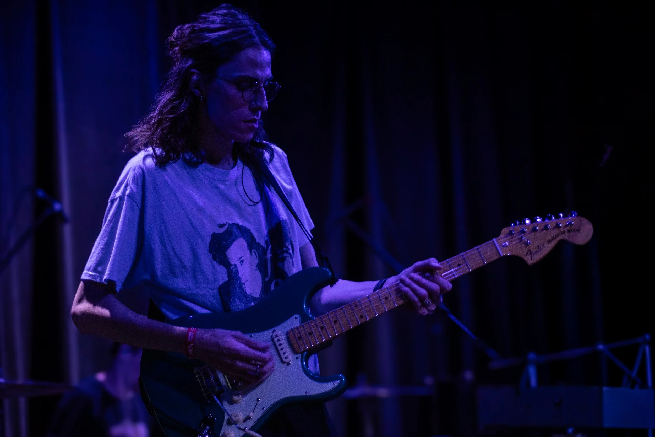 A woman playing an electric guitar on stage in a dimly lit environment, wearing glasses and a T-shirt with a graphic design.