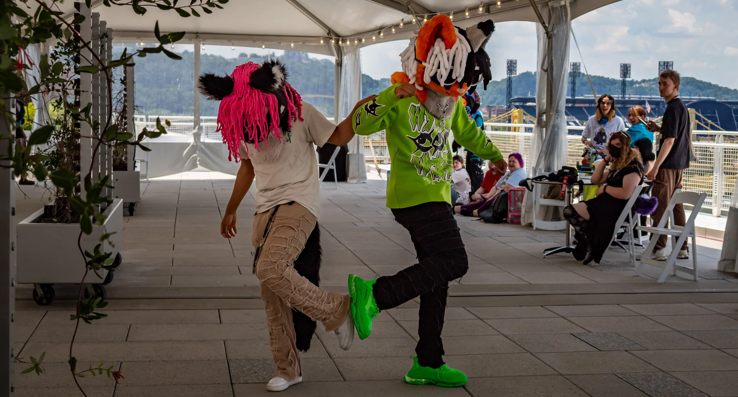Two people wearing colorful lion mascot costumes dancing together under a white tent with string lights, with a group of spectators seated and standing nearby.