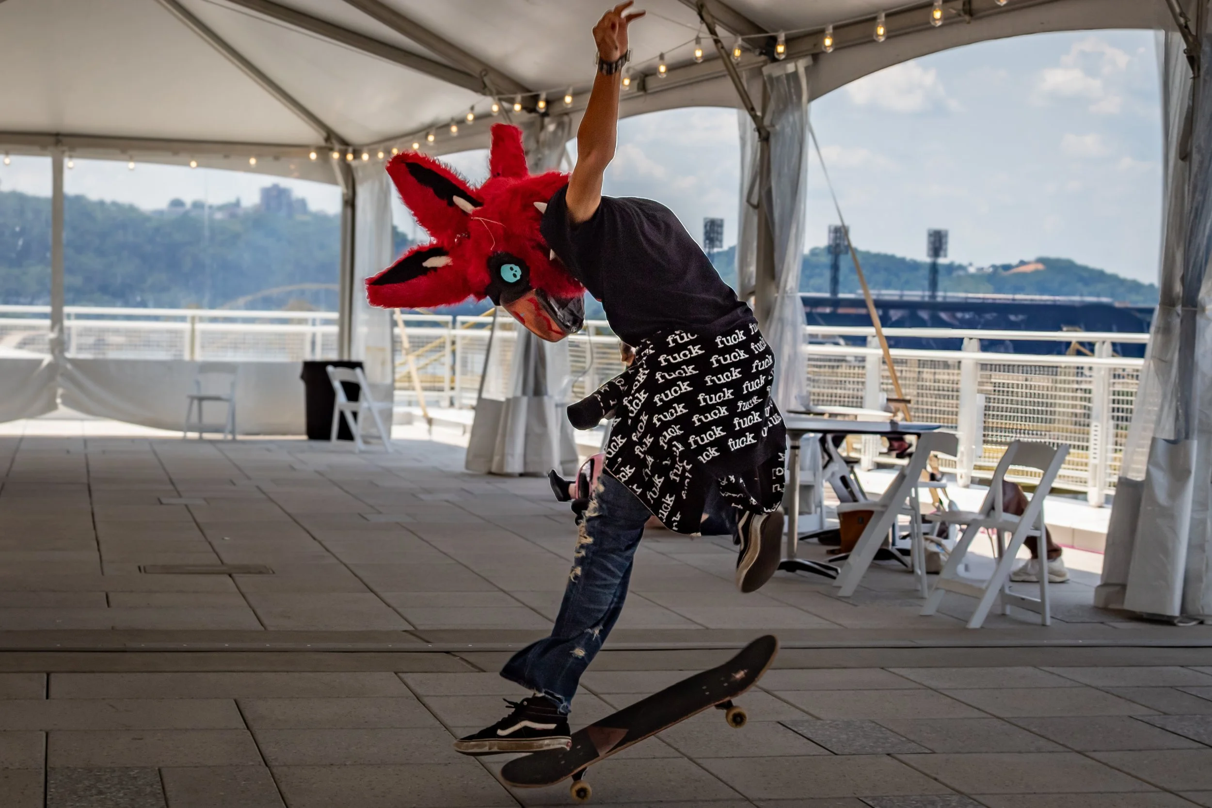 A person in a red fox costume with a large head and blue eye mask is skateboarding in a tented area with string lights and outdoor scenery in the background.