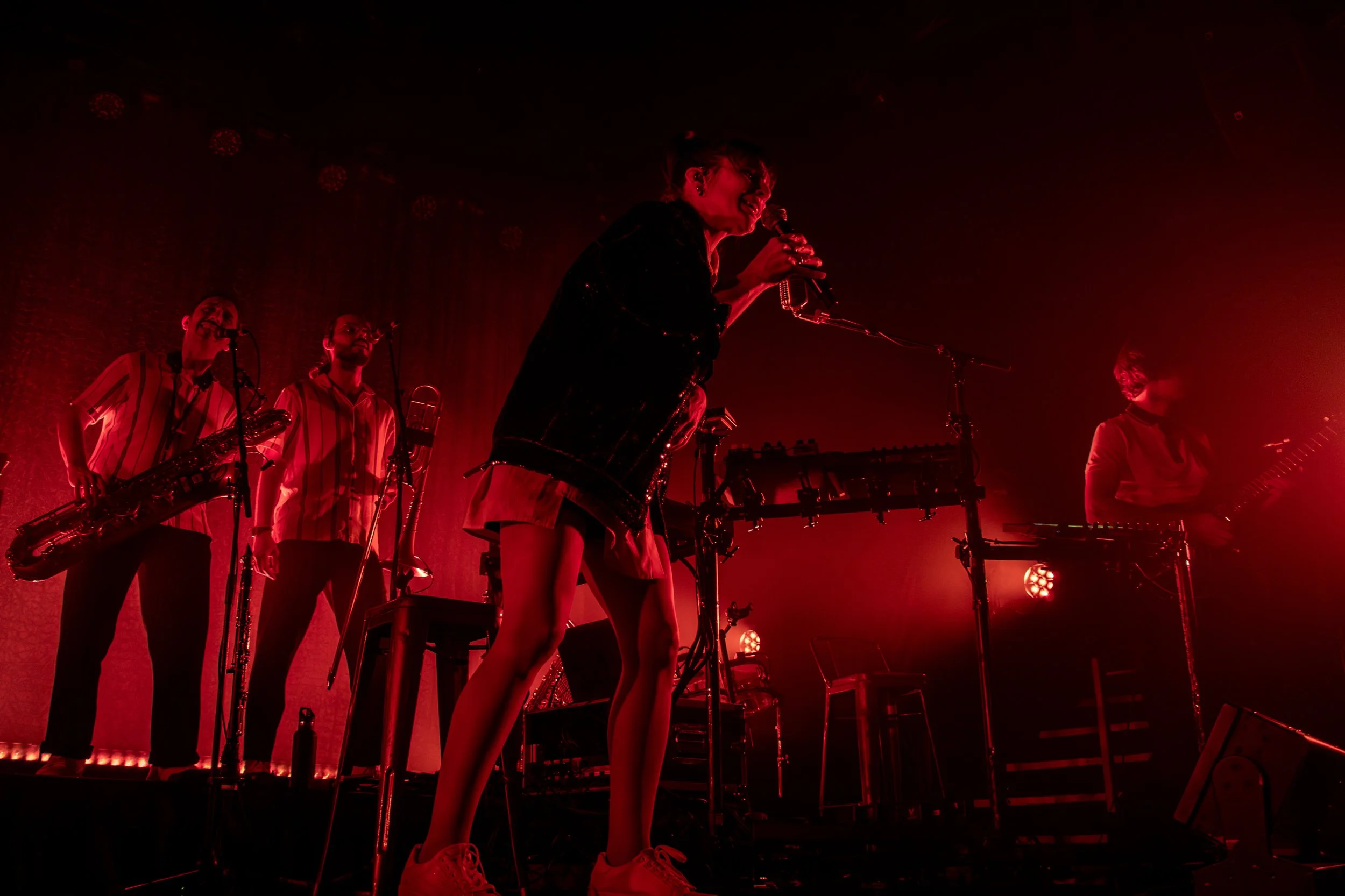 Band performing on stage under red lighting with musical instruments and electronic equipment.