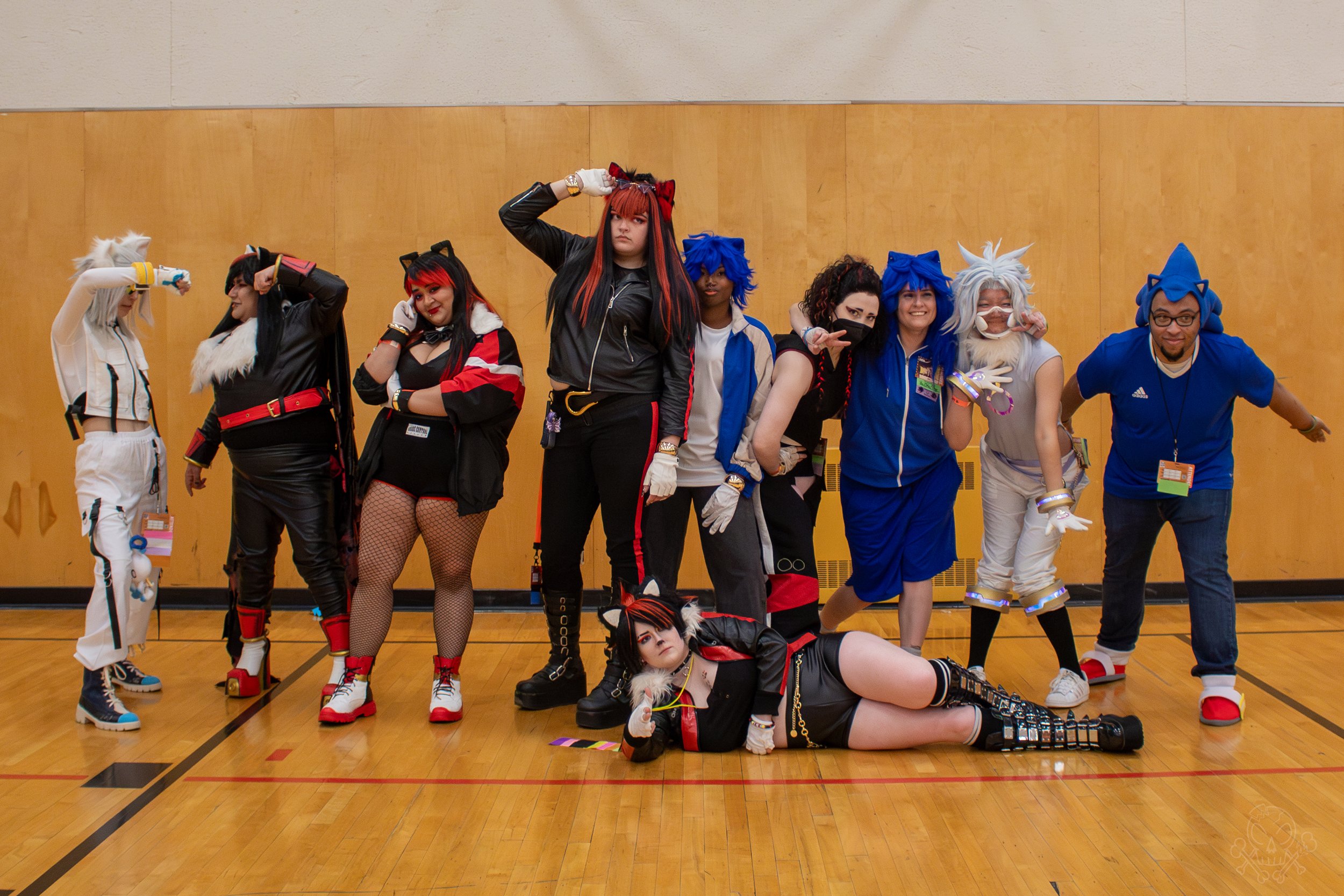 Group of people dressed in colorful anime or cosplay costumes, standing inside a gymnasium with a wood floor and beige wall.
