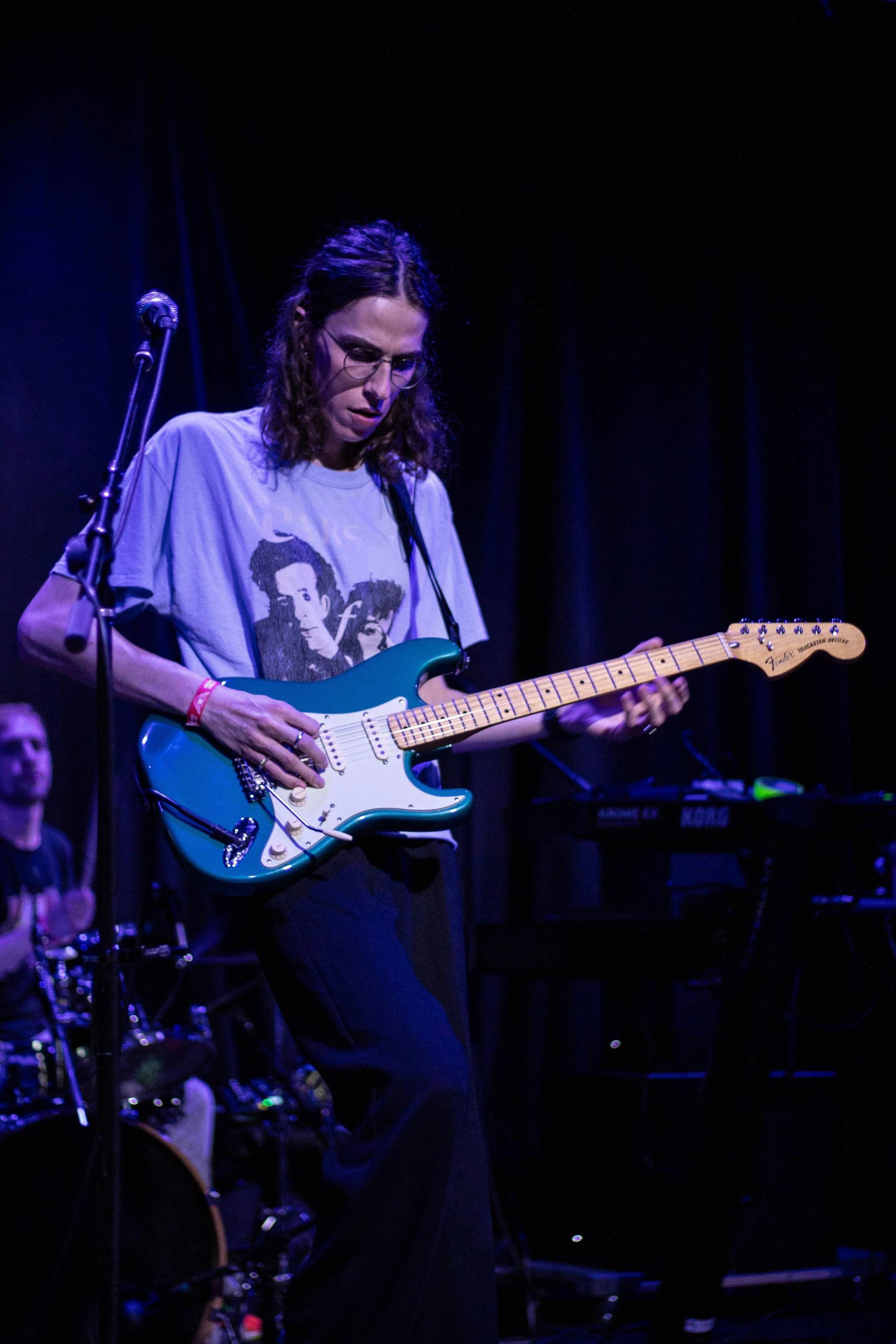 Person playing an electric guitar on stage, with keyboard and drums in background, dark stage lighting.
