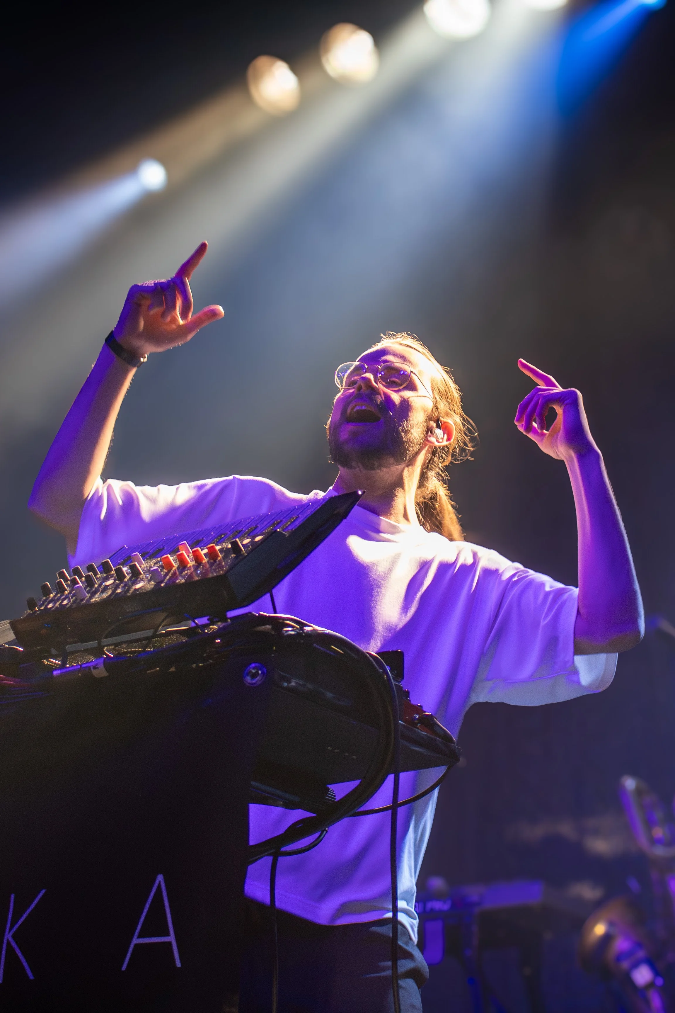 A DJ with glasses, long hair, and a beard is performing on stage with hands raised, surrounded by colorful stage lights.