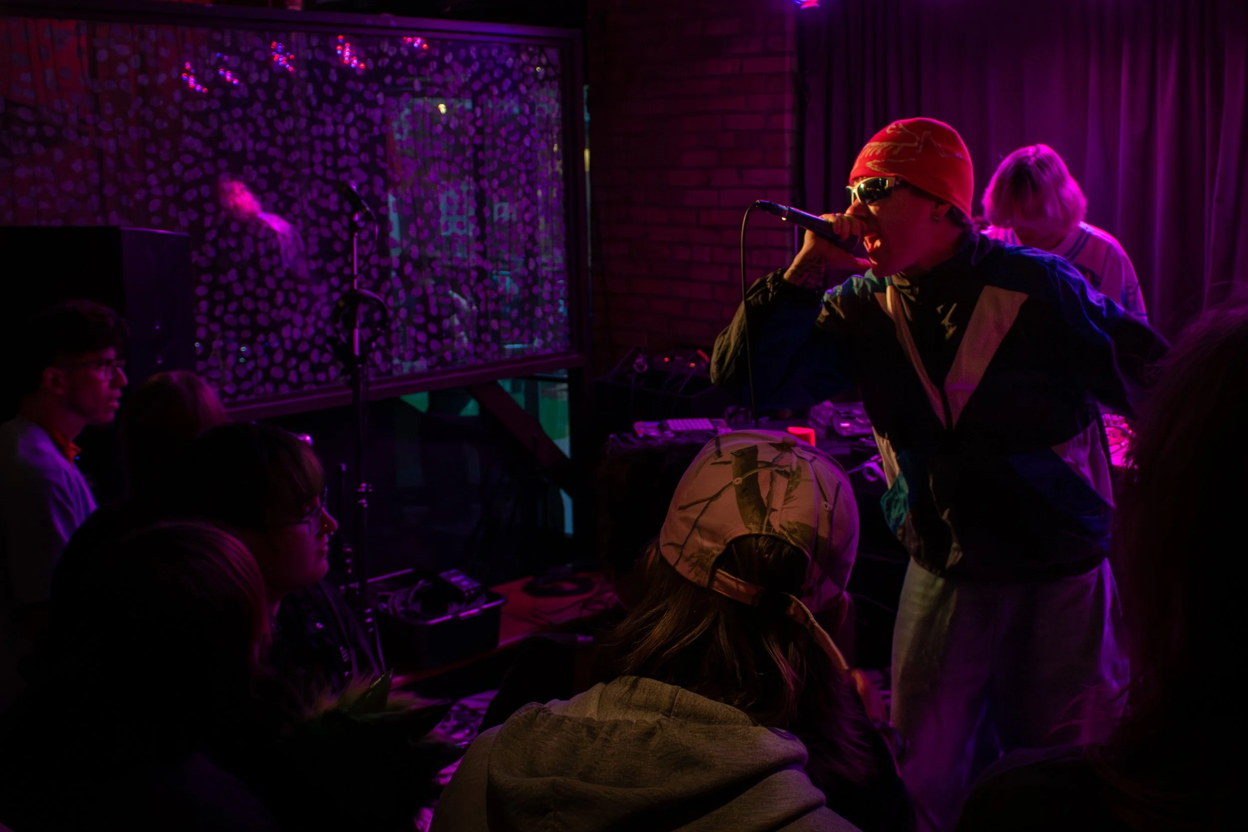 A performer singing into a microphone on stage during a live music event in a dimly lit venue with purple and pink lighting, while audience members watch.