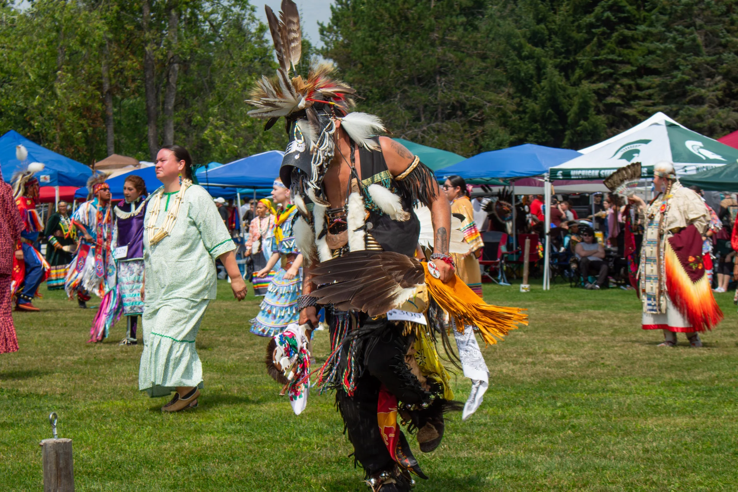 Native American dancers in traditional regalia performing at a cultural festival outdoors, with tents and trees in the background.