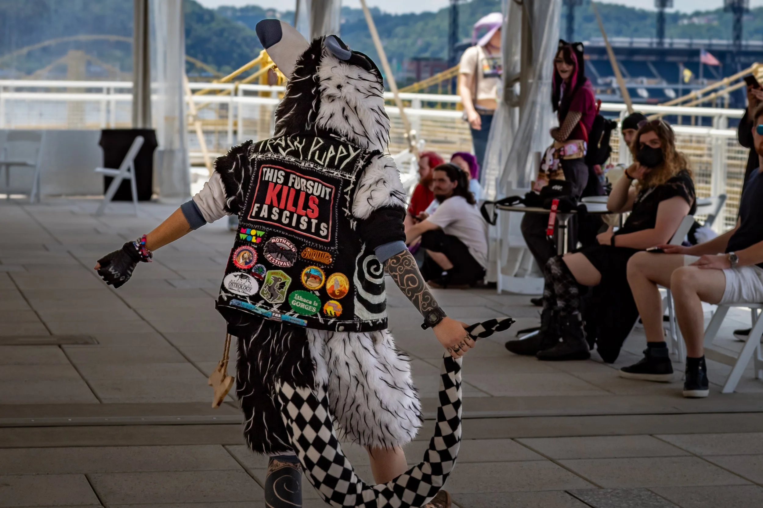 Person dressed in a furry wolf costume with patches and stickers on a vest, holding a black and white checkered tail, walking past seated observers at an outdoor event, with a shark costume headpiece and a painted arm.
