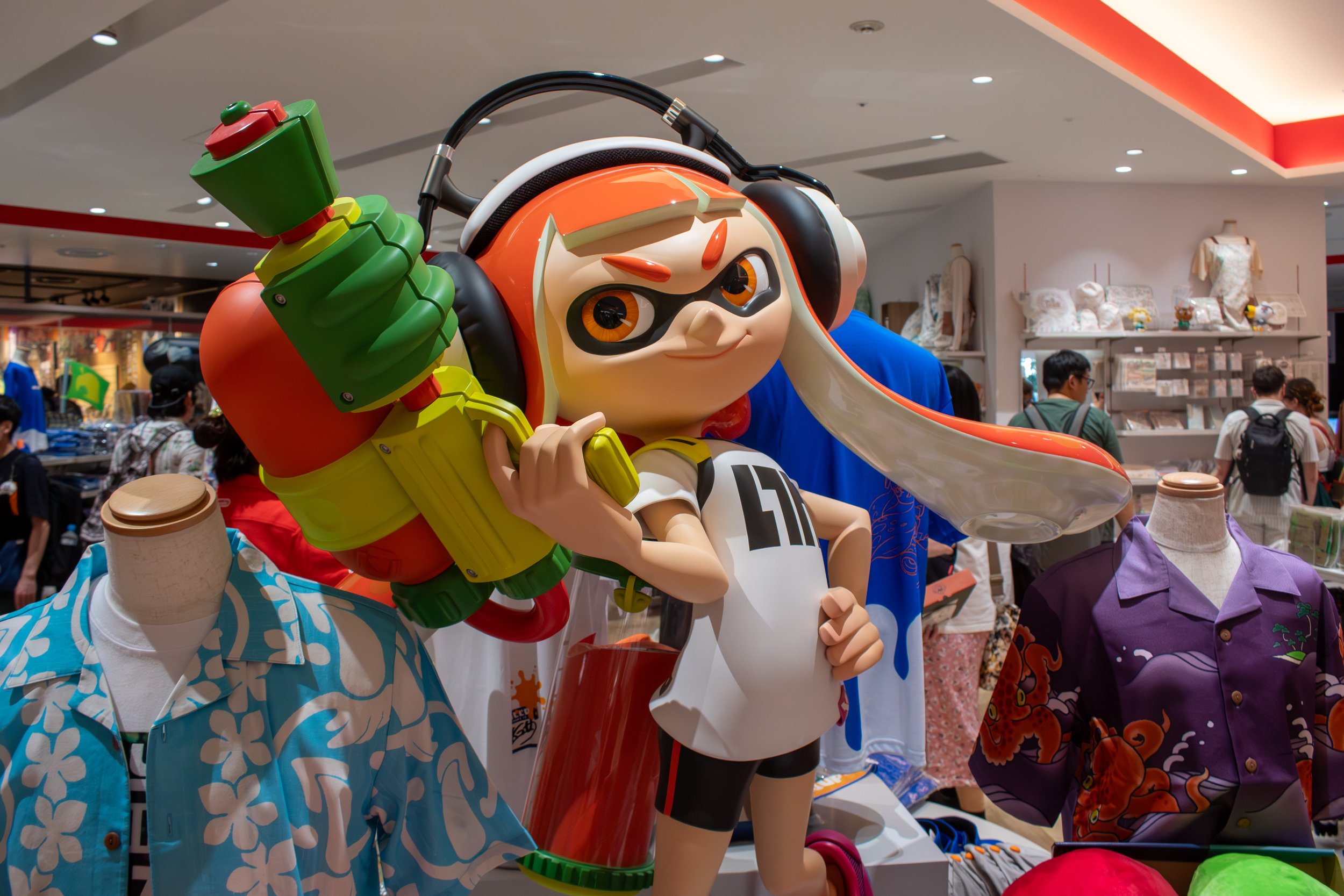 A statue of a character with orange and white hair, wearing a white jersey with the number 10, holding a large, colorful water gun, displayed in a store with mannequins dressed in Hawaiian shirts and shoppers in the background.