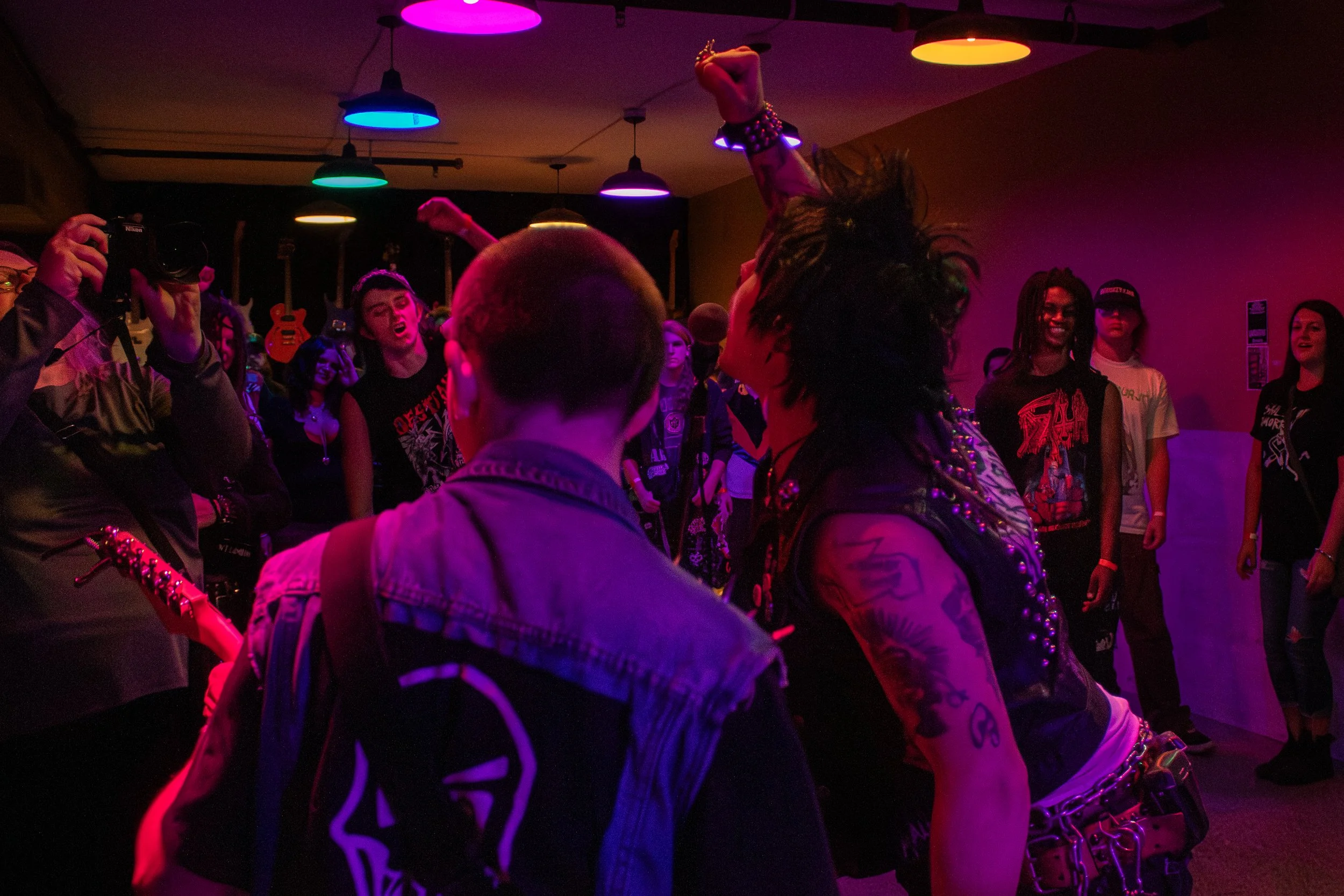 A punk rock concert in a dimly lit room with colorful ceiling lights. A woman with tattoos and dark hair, wearing a leather vest, is dancing with her arm raised. Several people are watching and taking photos, some wearing band t-shirts, with guitars 