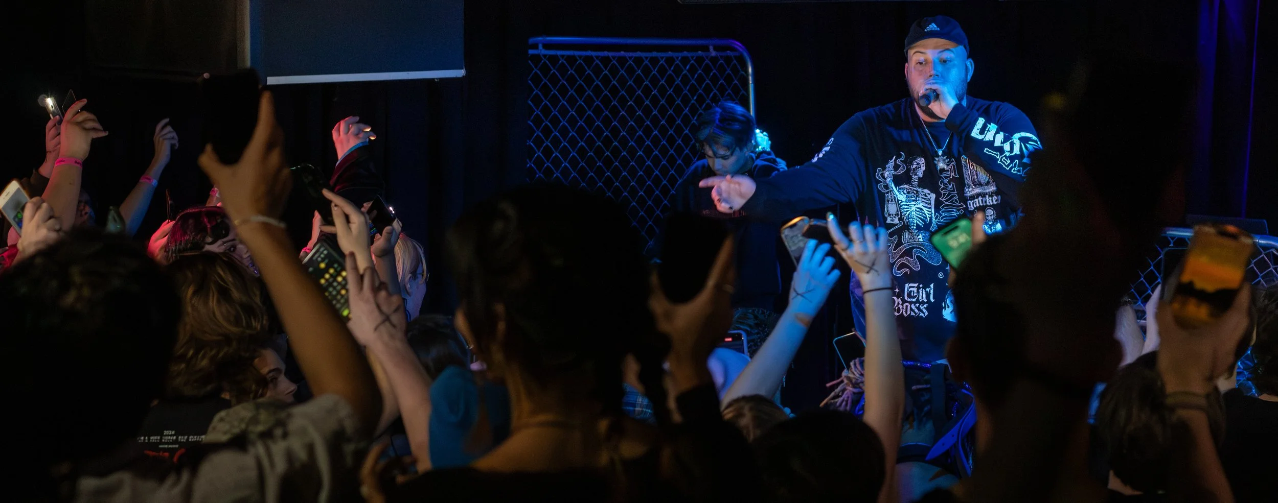 Male performer wearing a black cap and black long-sleeve shirt with graphics, holding a microphone, on stage with audience taking photos