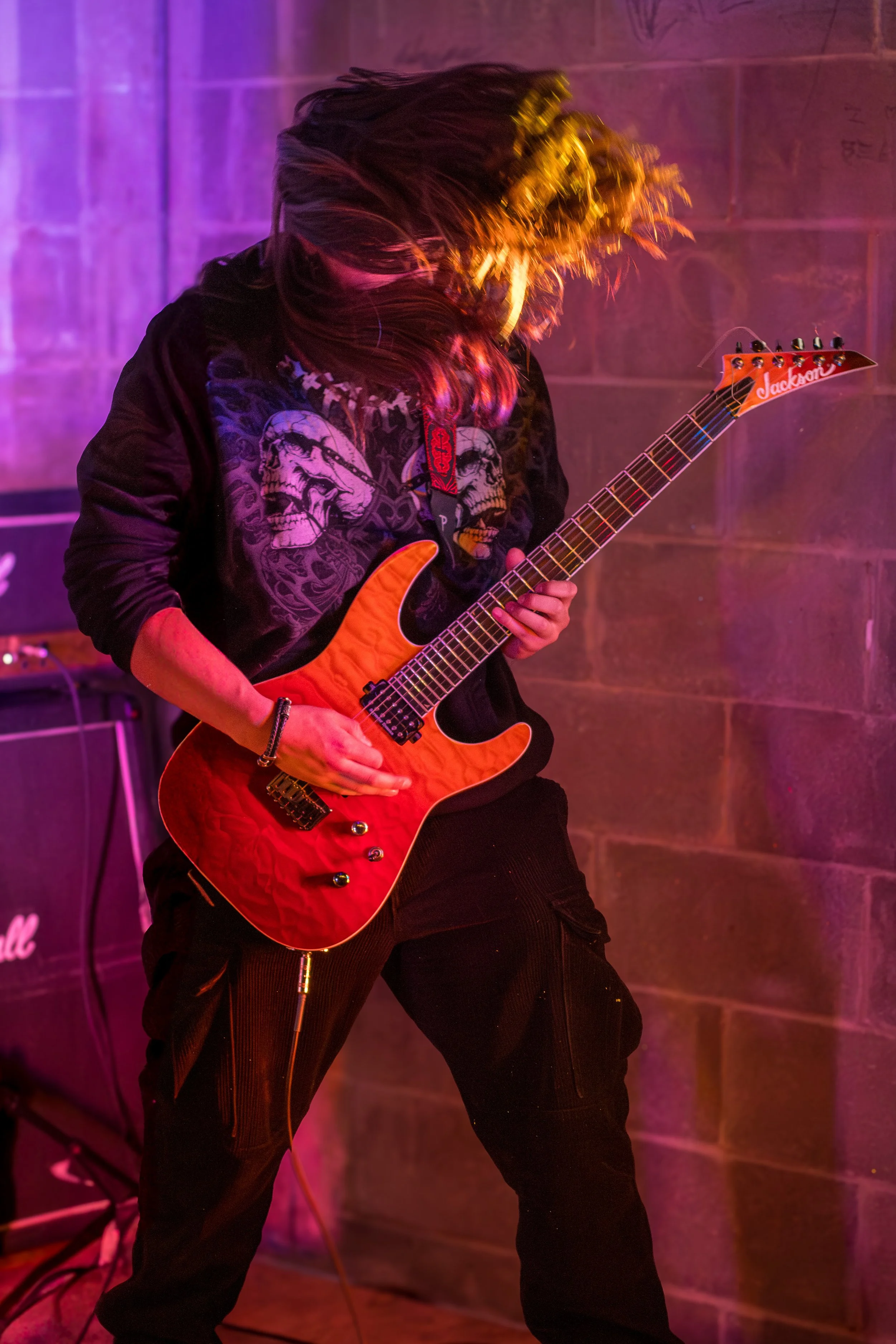 A person playing an electric guitar with long hair swinging, wearing a black hoodie with skull graphics, in a room with colorful lighting and a brick wall background.