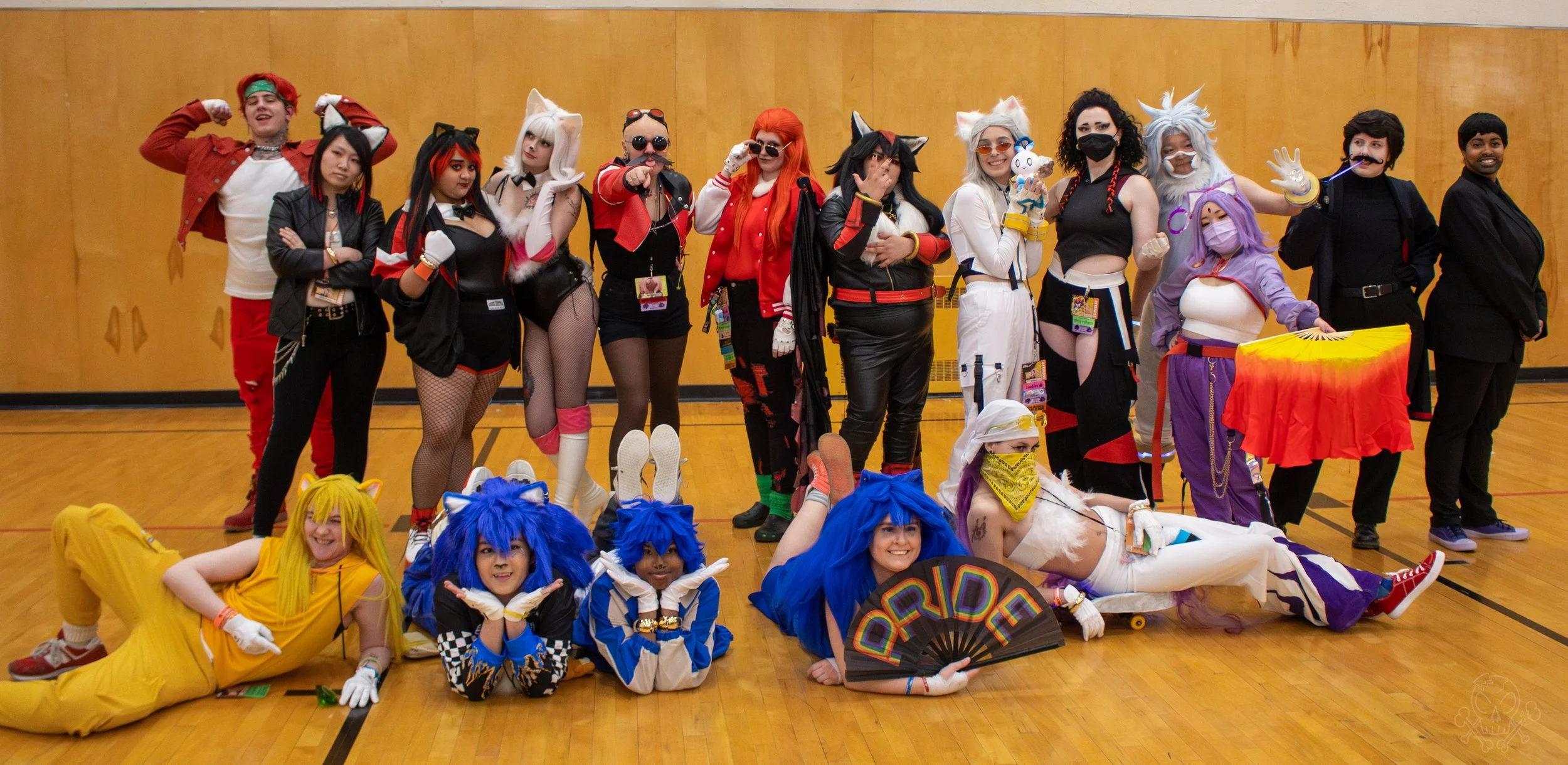 Group of people in colorful, elaborate costumes at a cosplay event, posing together in a gymnasium with wooden flooring and a tan wall.