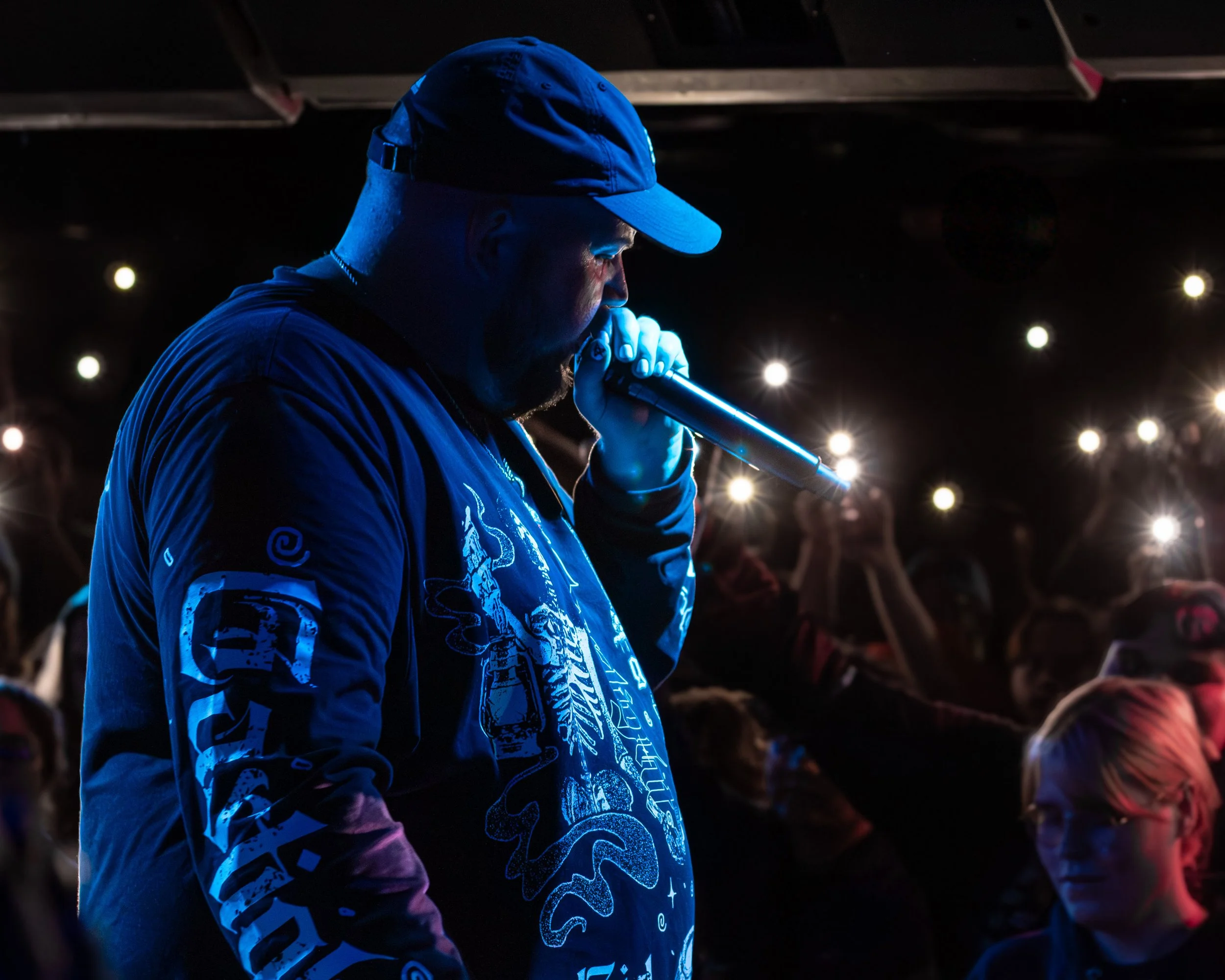 A male performer or rapper on stage, holding a microphone close to his mouth, with a dark background and audience members holding up smartphones with illuminated flashlights.