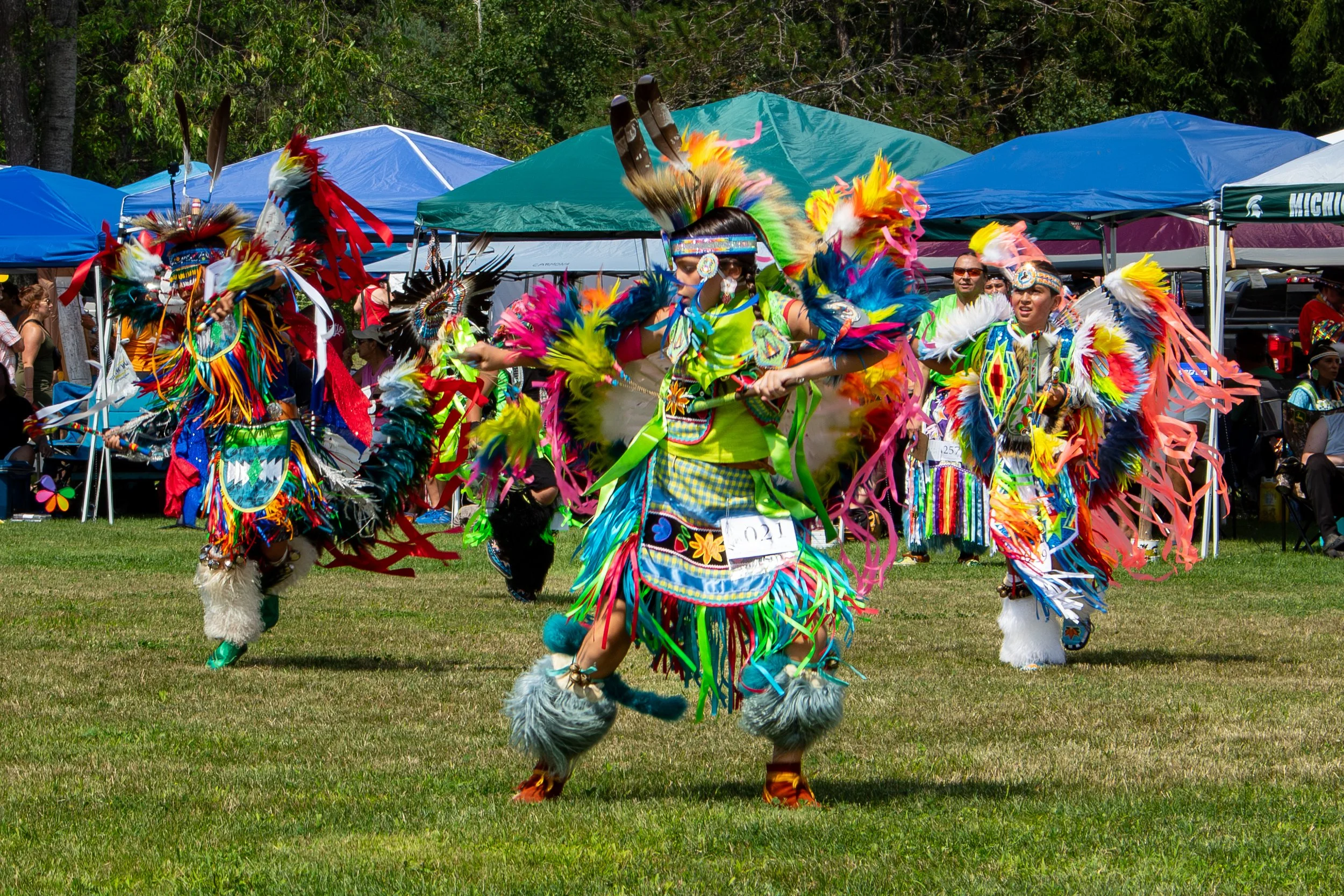 Native American dancers in colorful traditional attire performing at a powwow, with blue and green tents in the background.