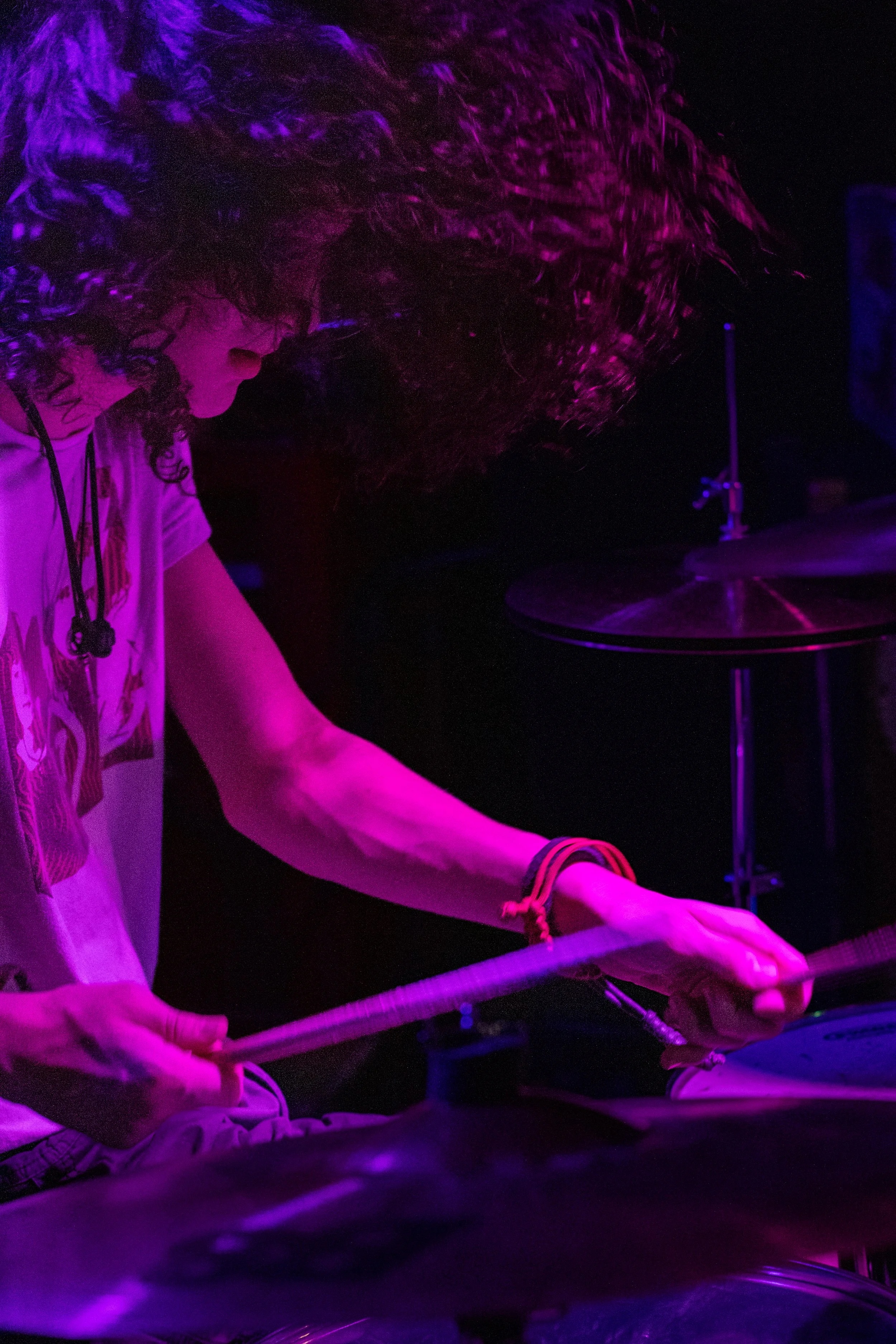 A person with long curly hair is playing the drums under purple lighting, with a drumstick in one hand, wearing a graphic t-shirt and multiple bracelets.
