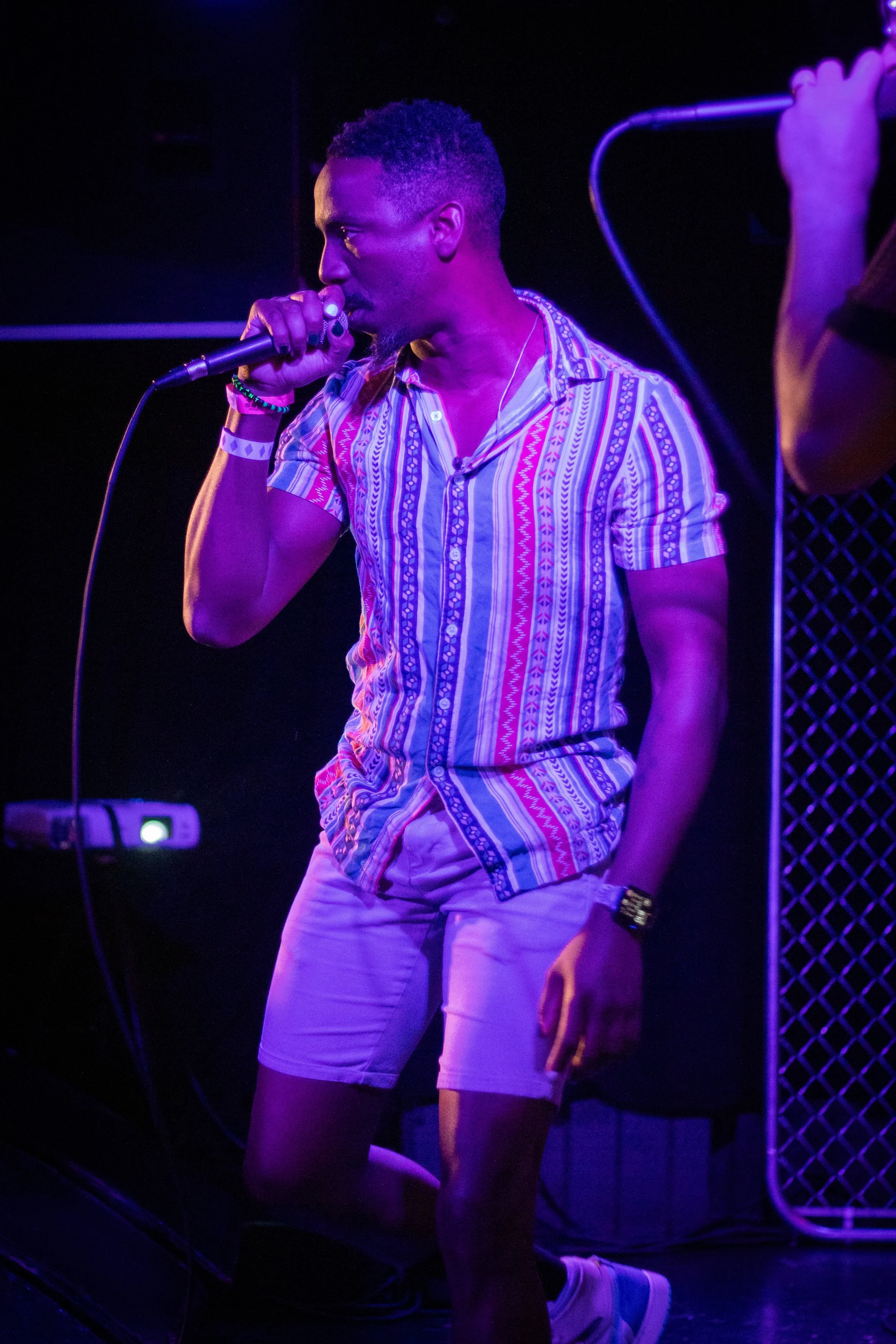 Man singing into a microphone on stage, wearing colorful striped shirt, shorts, and sneakers, with purple lighting.
