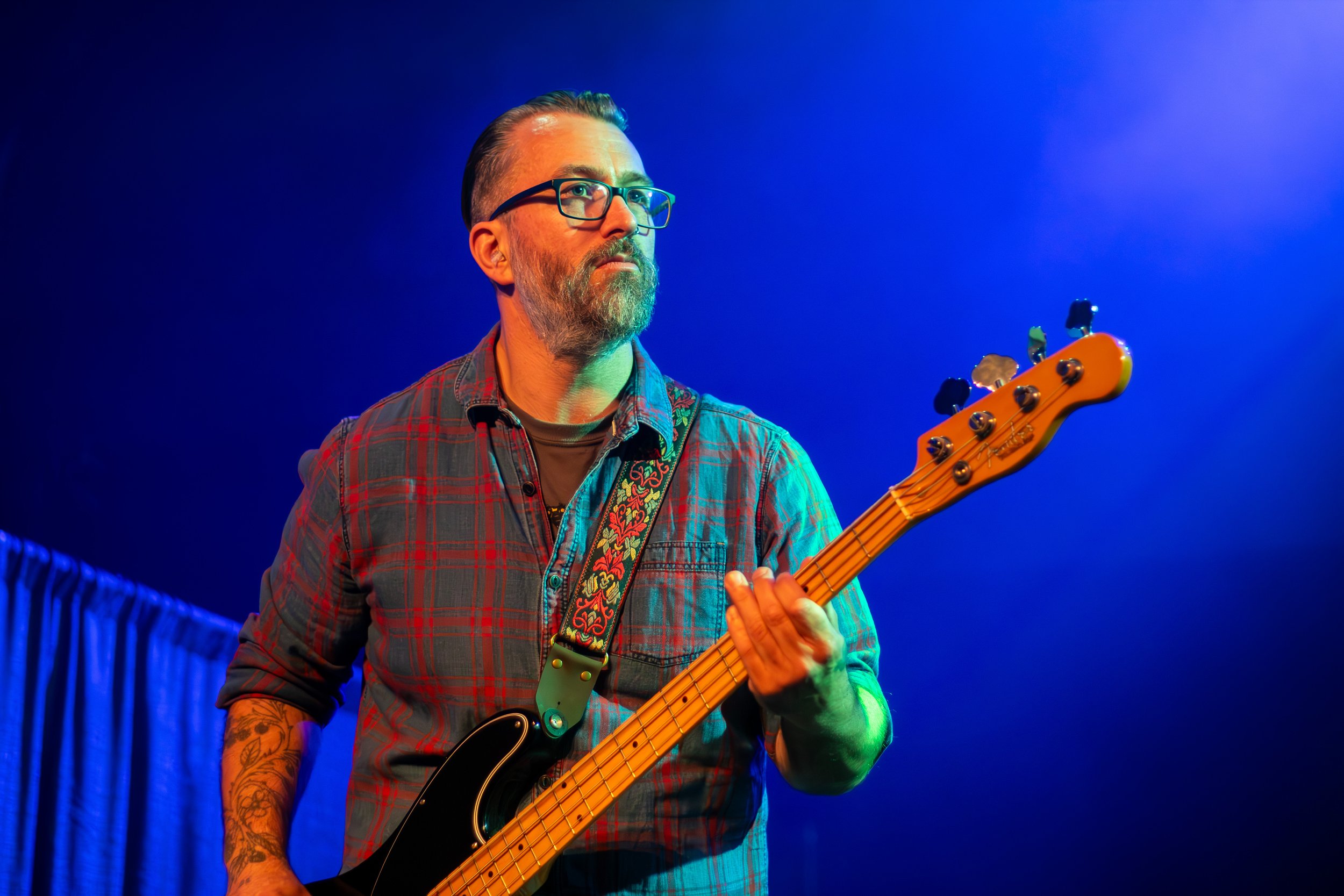 Man with glasses and a beard playing an electric bass guitar on stage under blue lighting.
