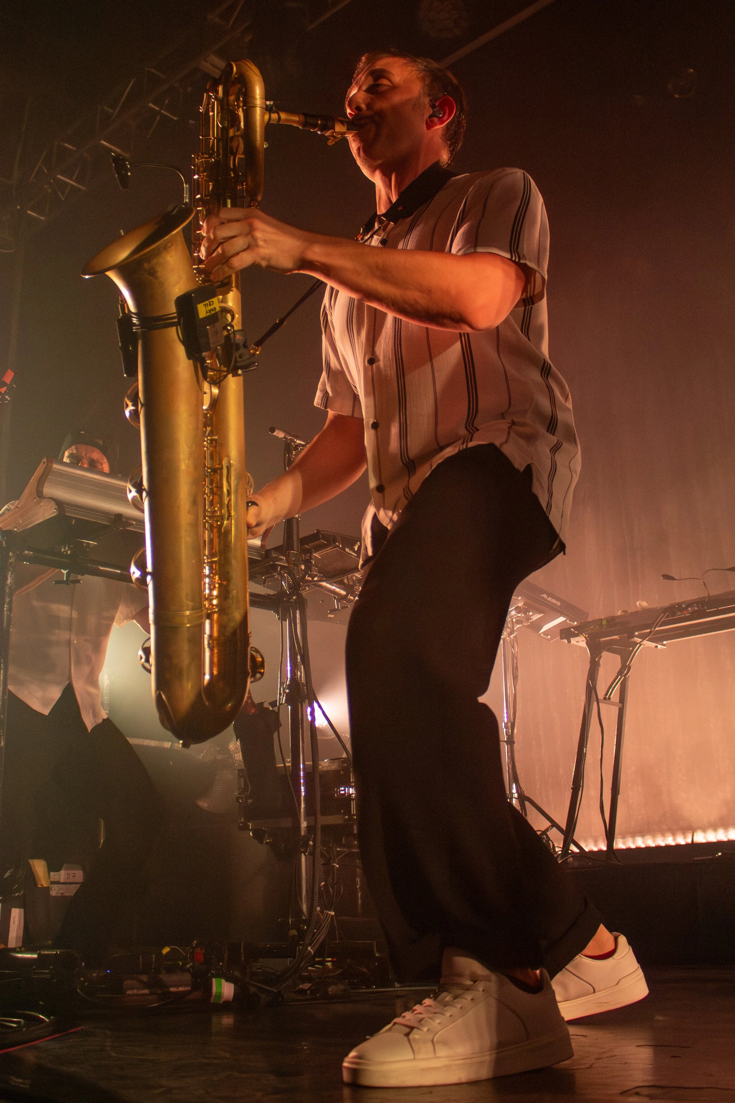 A musician playing a saxophone on stage during a concert, with stage lighting and electronic music equipment in the background.