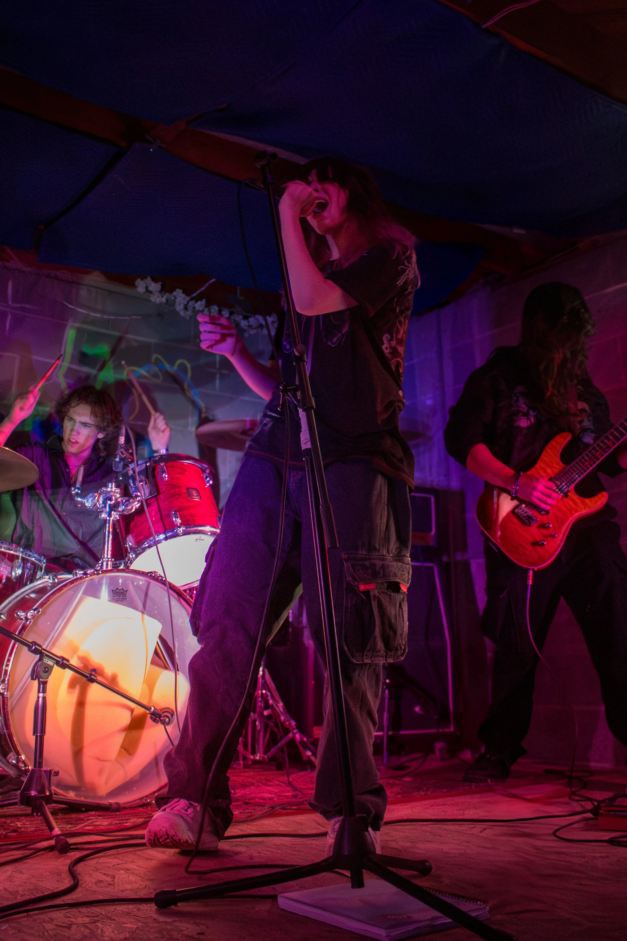 A band performs on stage with a singer, drummer, and guitarist under colorful stage lights.