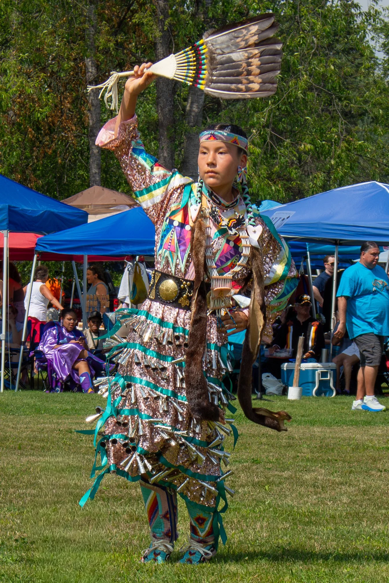 Native American dancer in traditional regalia holding a feather fan at a cultural event with spectators and tents in the background.