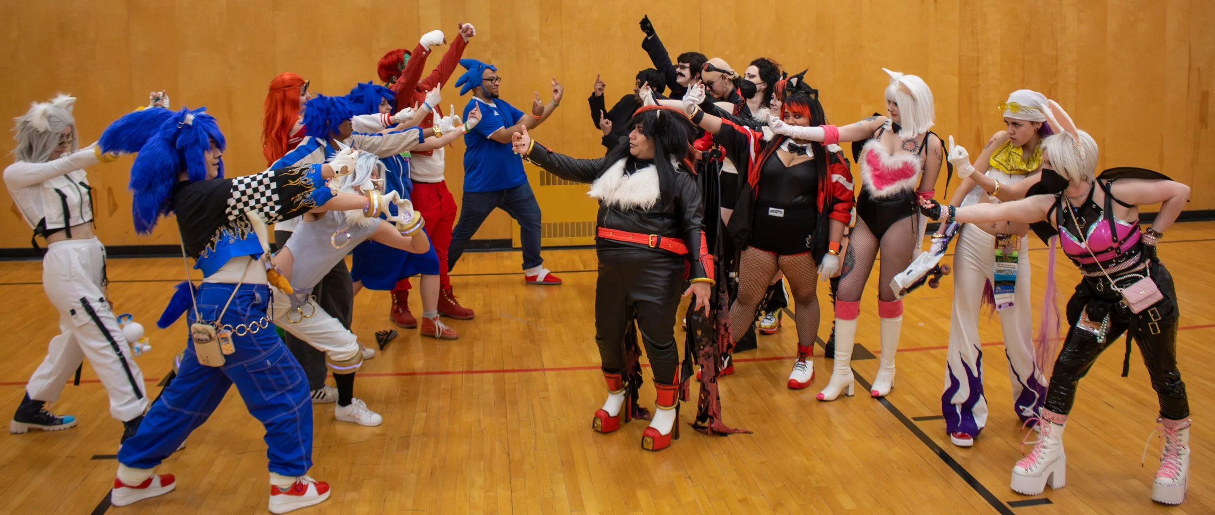 People dressed in colorful, playful costumes and wigs, gathered in a gymnasium, posing in two lines for a group photo.