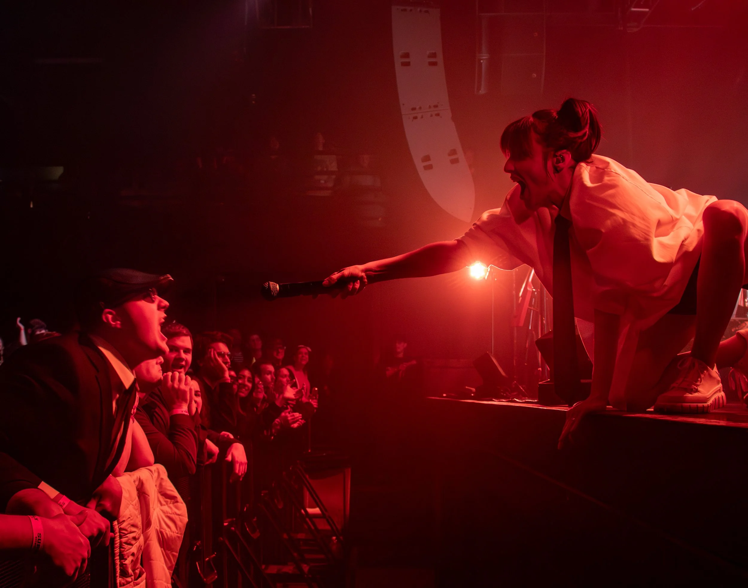 Performer in white shirt and black tie on stage reaching out with microphone to fan in audience, group of fans cheering and watching.