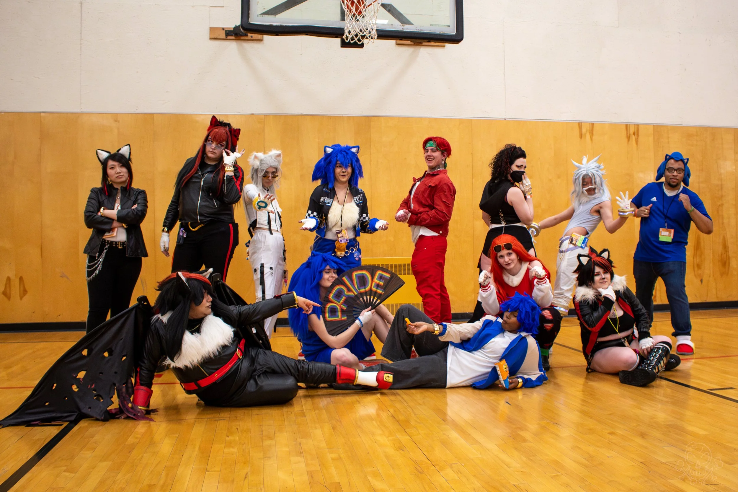 Group of people in colorful cosplay costumes with animal ears and tails, posing together in a gymnasium with a basketball hoop above.