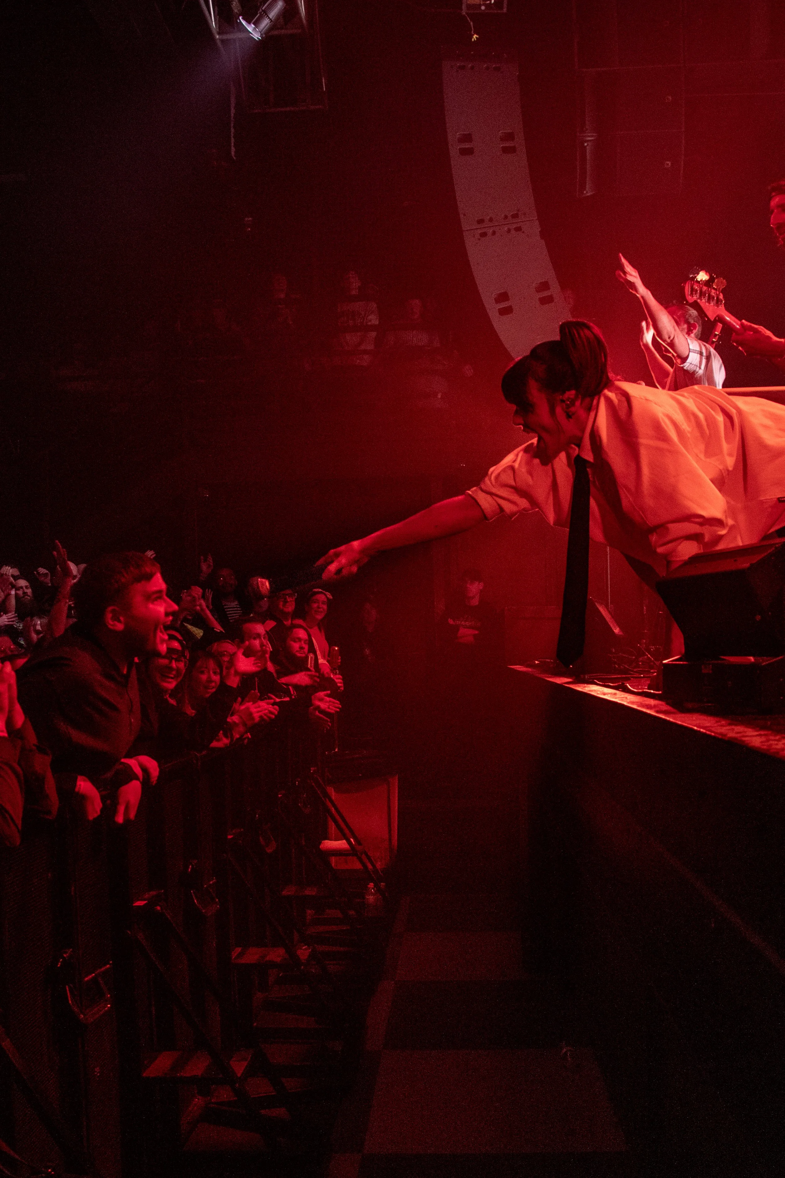Performer on stage reaching out to enthusiastic audience in a concert venue with red lighting.