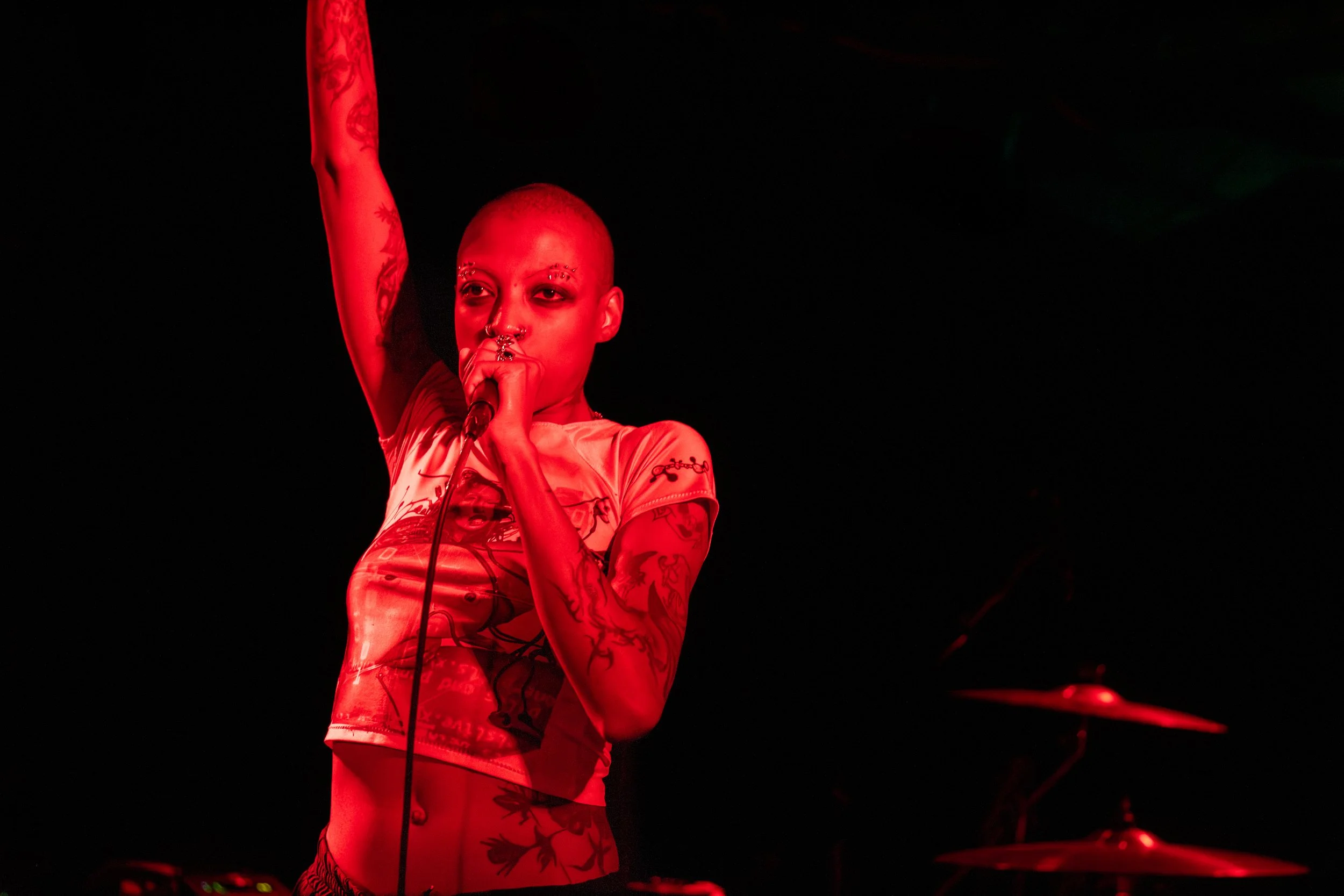 A person with short hair and tattoos performing on stage, holding a microphone near their mouth, illuminated by red lighting, with a dark background.