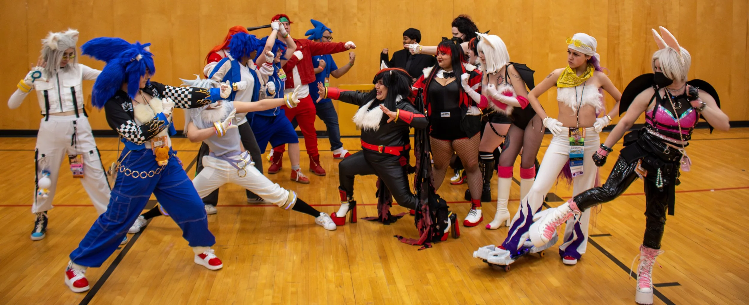 Group of people dressed in colorful costumes, costumes resemble anthropomorphic characters, posing on a wooden gymnasium floor.