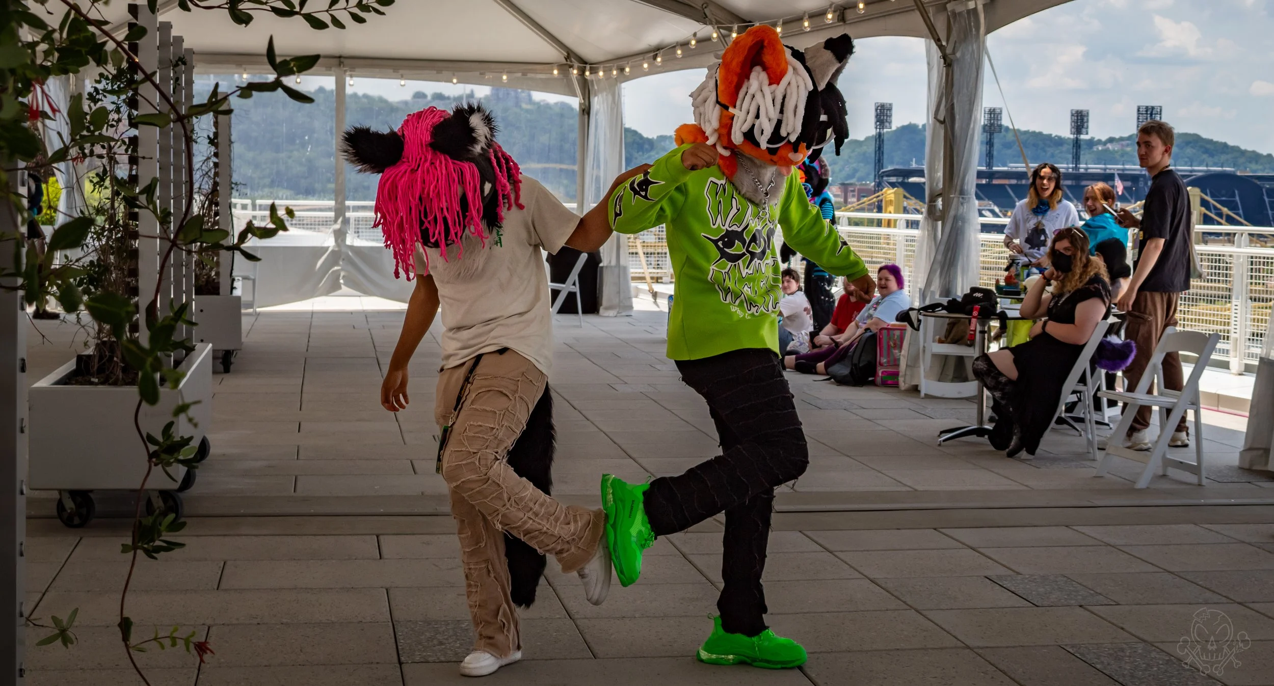Two people wearing colorful animal mascot costumes dancing together at an outdoor event, with an audience in the background.