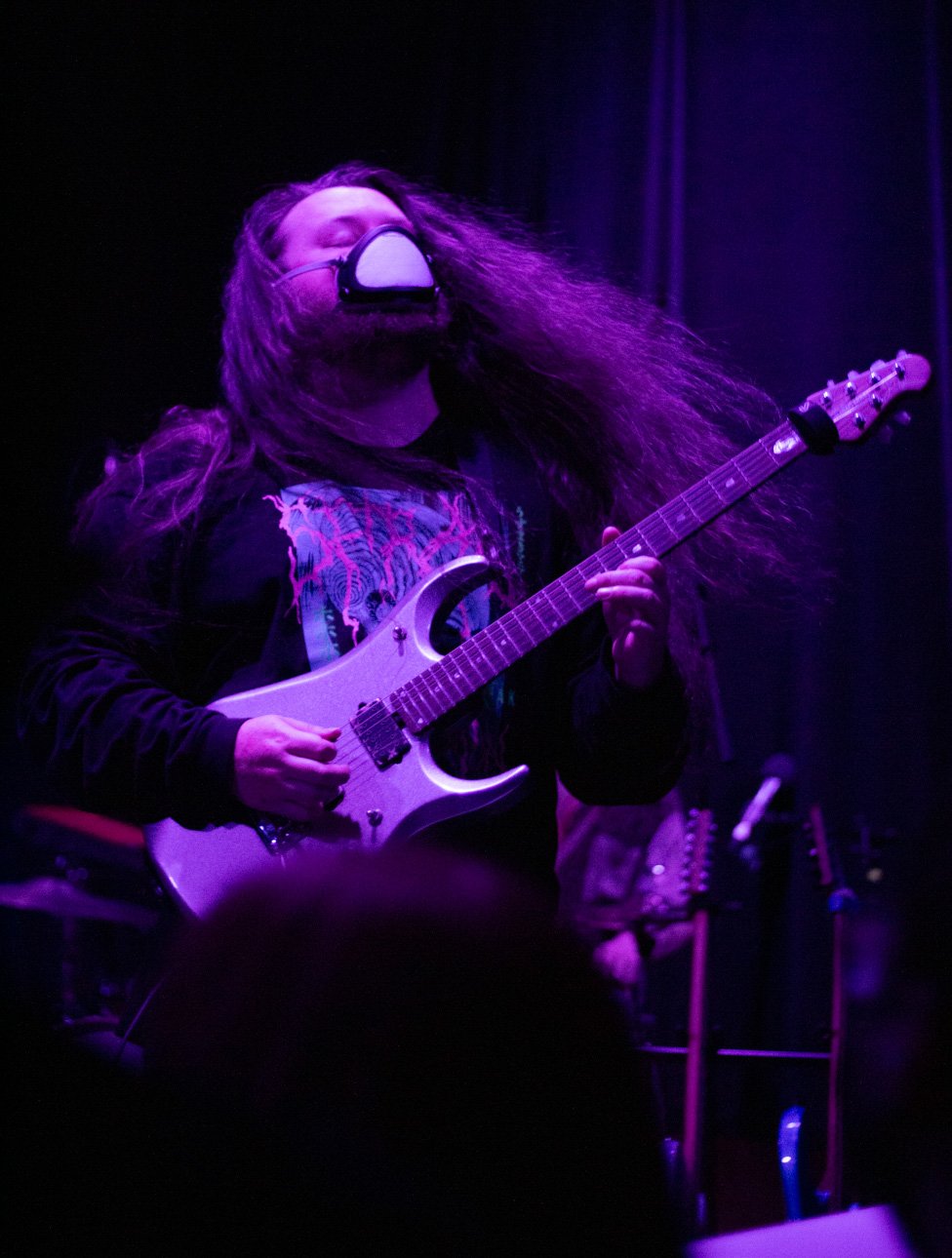 A person with long hair wearing a face mask and playing an electric guitar on stage under purple lighting, with a drum set in the background.