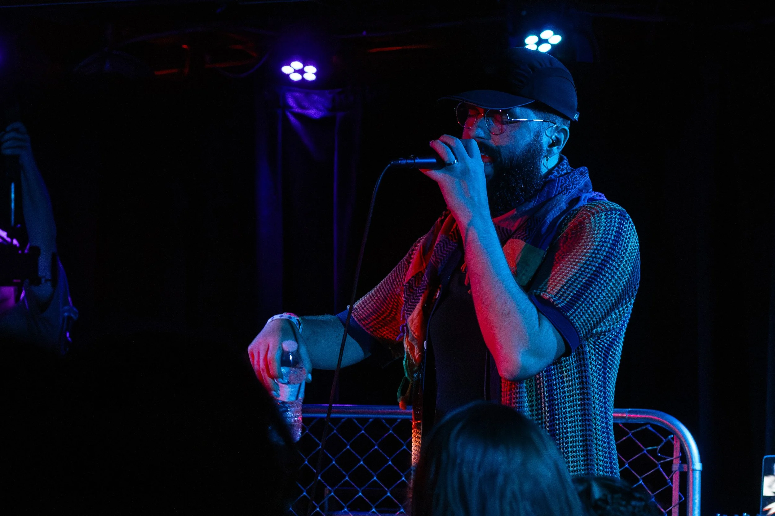 A man with a beard, glasses, a black cap, and a colorful shirt singing into a microphone on stage, holding a water bottle in his other hand, with stage lights illuminating in blue and purple
